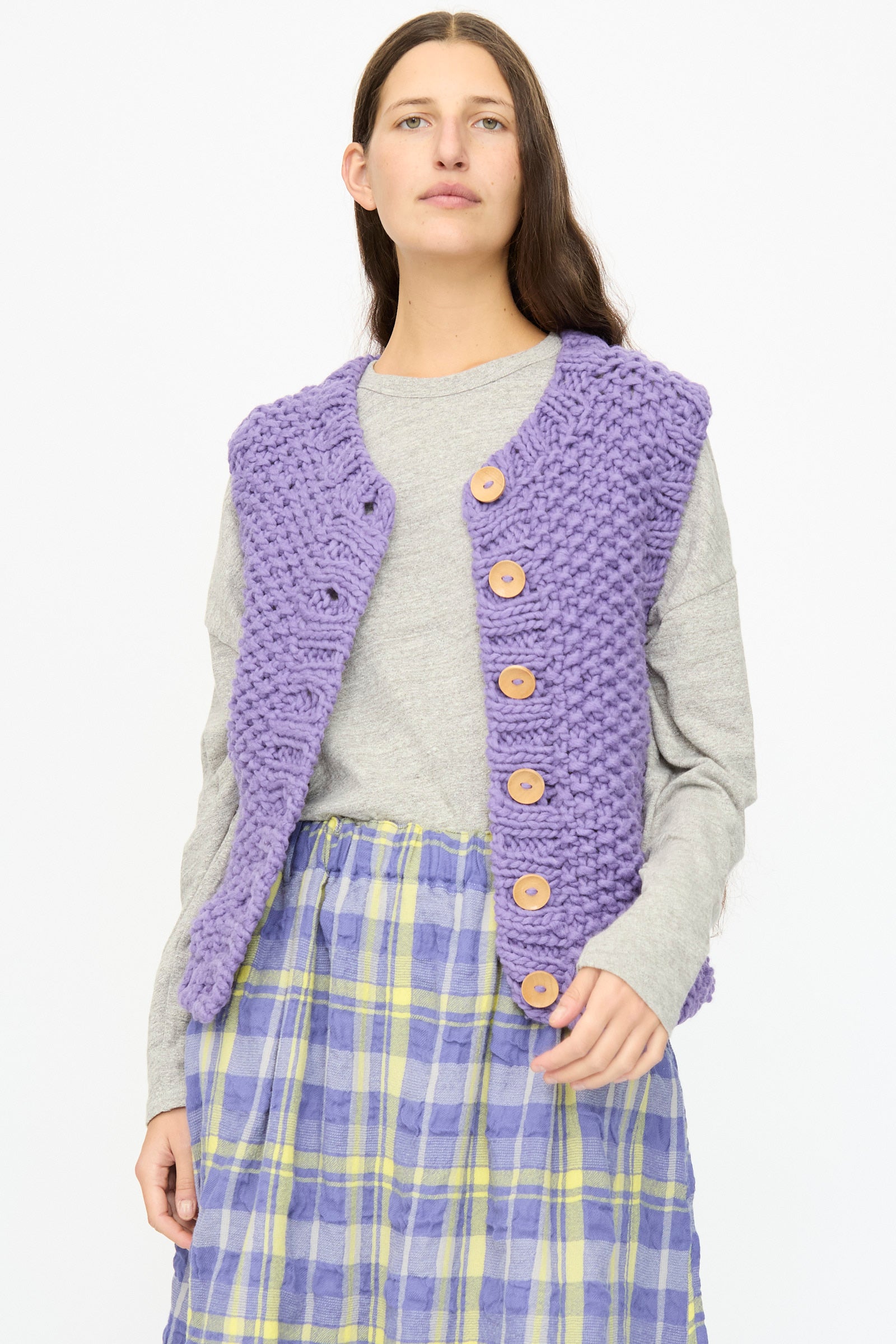 A person wears the Ichi Antiquités Wool Peru Hand-knit Vest in Purple over a gray long-sleeve shirt and a purple plaid skirt, standing facing forward.