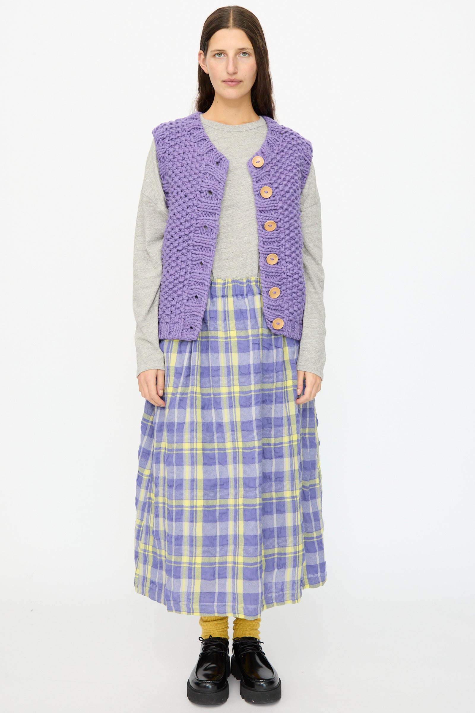 A woman faces forward against a white background wearing the Ichi Antiquités Wool Peru Hand-knit Vest in Purple, paired with a gray long-sleeve shirt, purple and yellow plaid skirt, mustard socks, and black shoes.