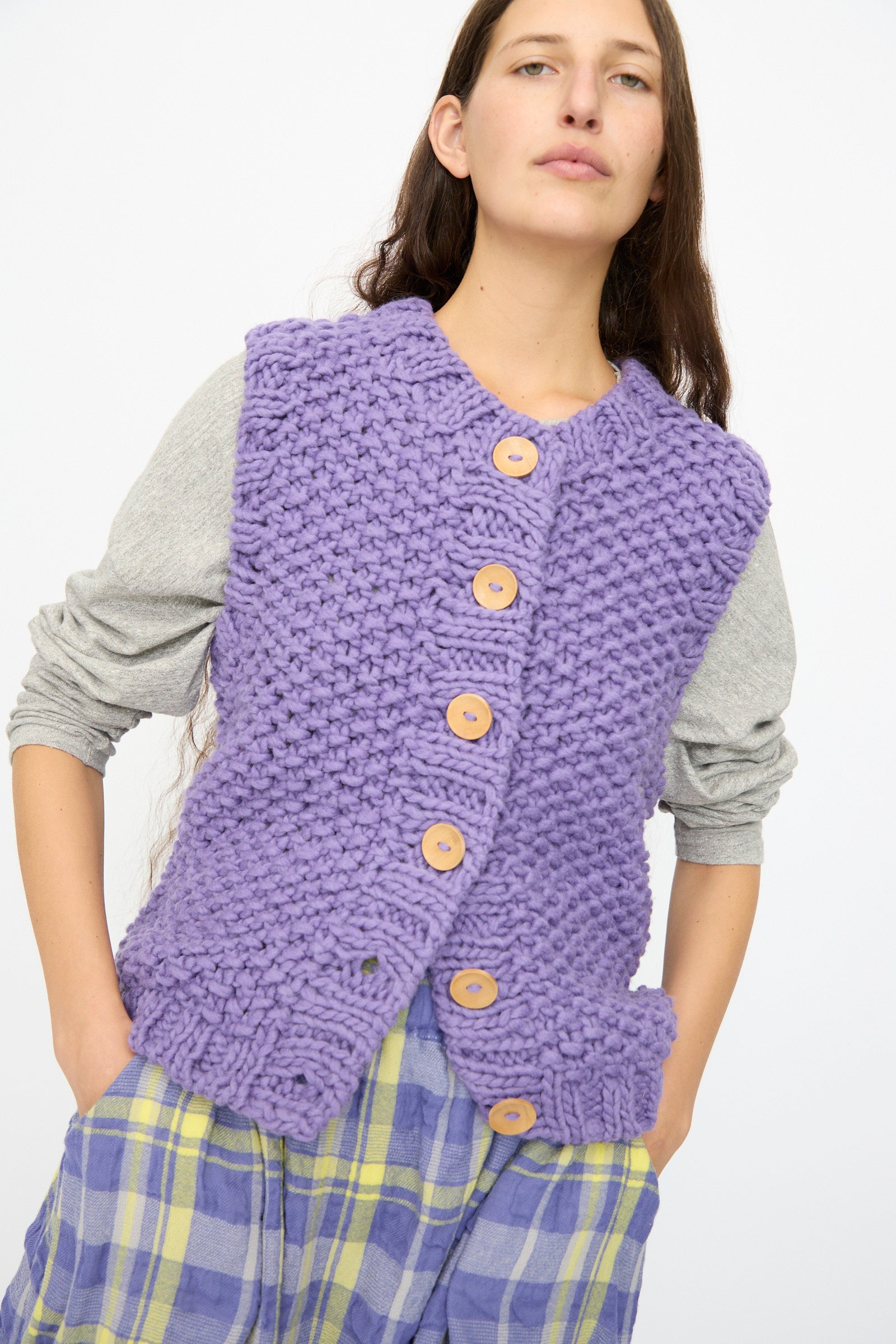 A person wears the Ichi Antiquités Wool Peru Hand-knit Vest in Purple over a grey long-sleeve shirt and a purple and yellow plaid skirt.
