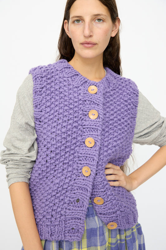 A woman stands with one hand on her hip, wearing the Ichi Antiquités Wool Peru Hand-knit Vest in Purple with large wooden buttons over a gray long-sleeve top and a plaid skirt.