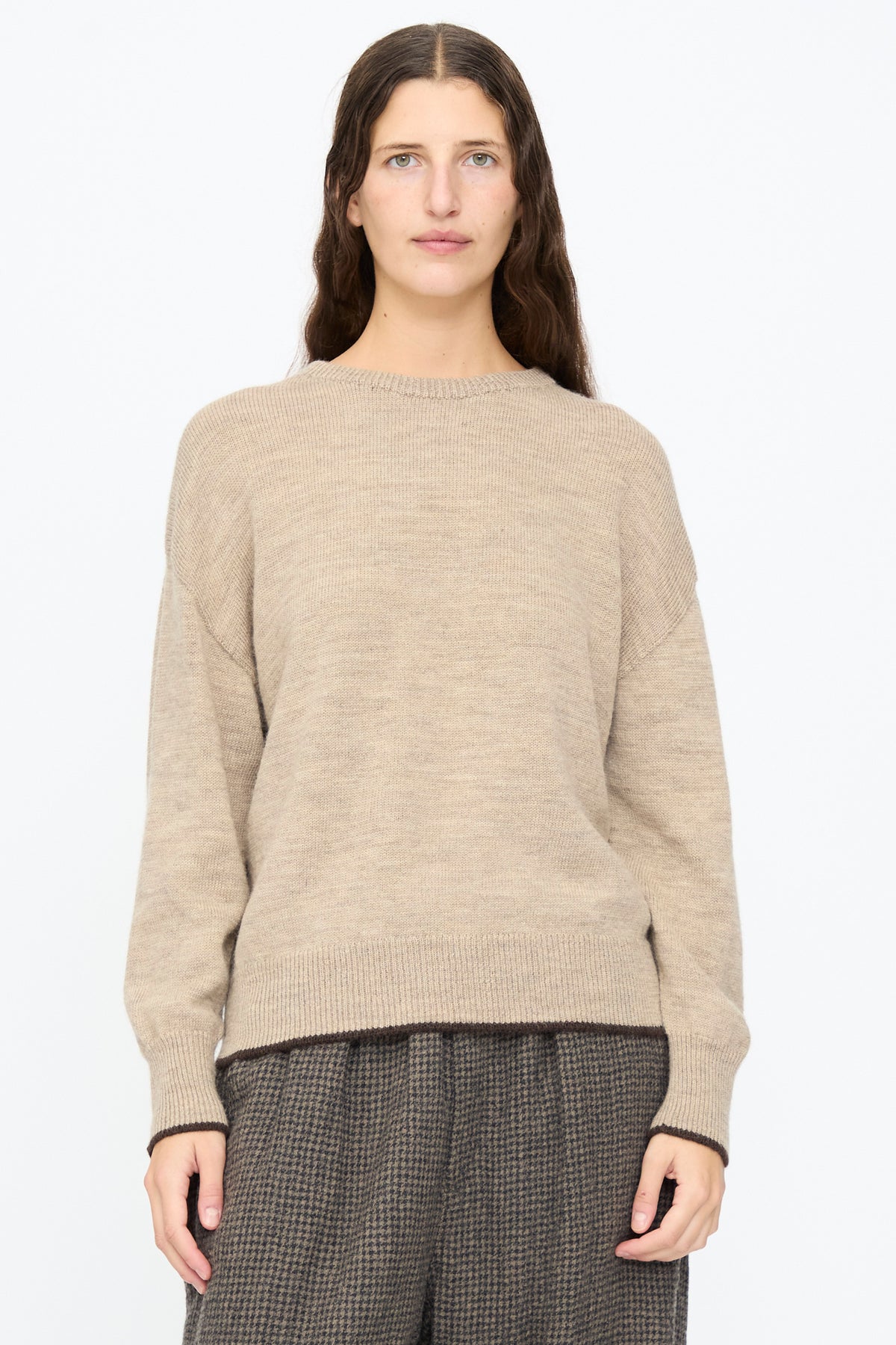 A person wearing an Ichi Antiquités Wool Pullover in Beige and Brown, featuring dark brown trim at the hem and cuffs, paired with dark patterned trousers.