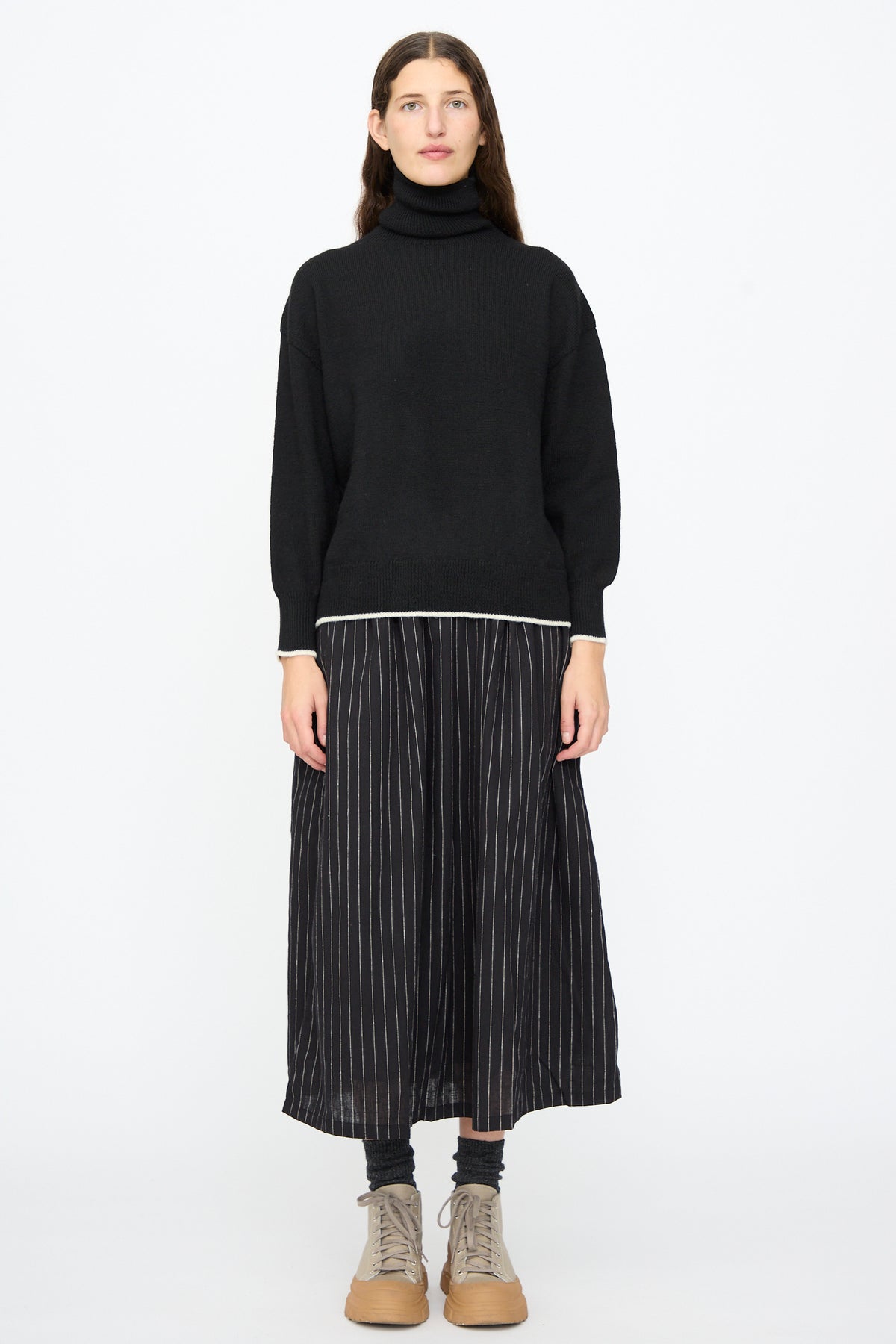A person wears an Ichi Antiquités Wool Pullover in Black and White paired with a black pinstriped midi skirt and grey lace-up boots.