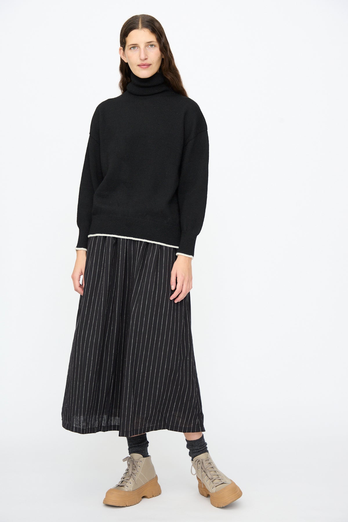 A person wearing an Ichi Antiquités Wool Pullover in Black and White paired with a black pinstriped midi skirt, gray socks, and tan lace-up boots.
