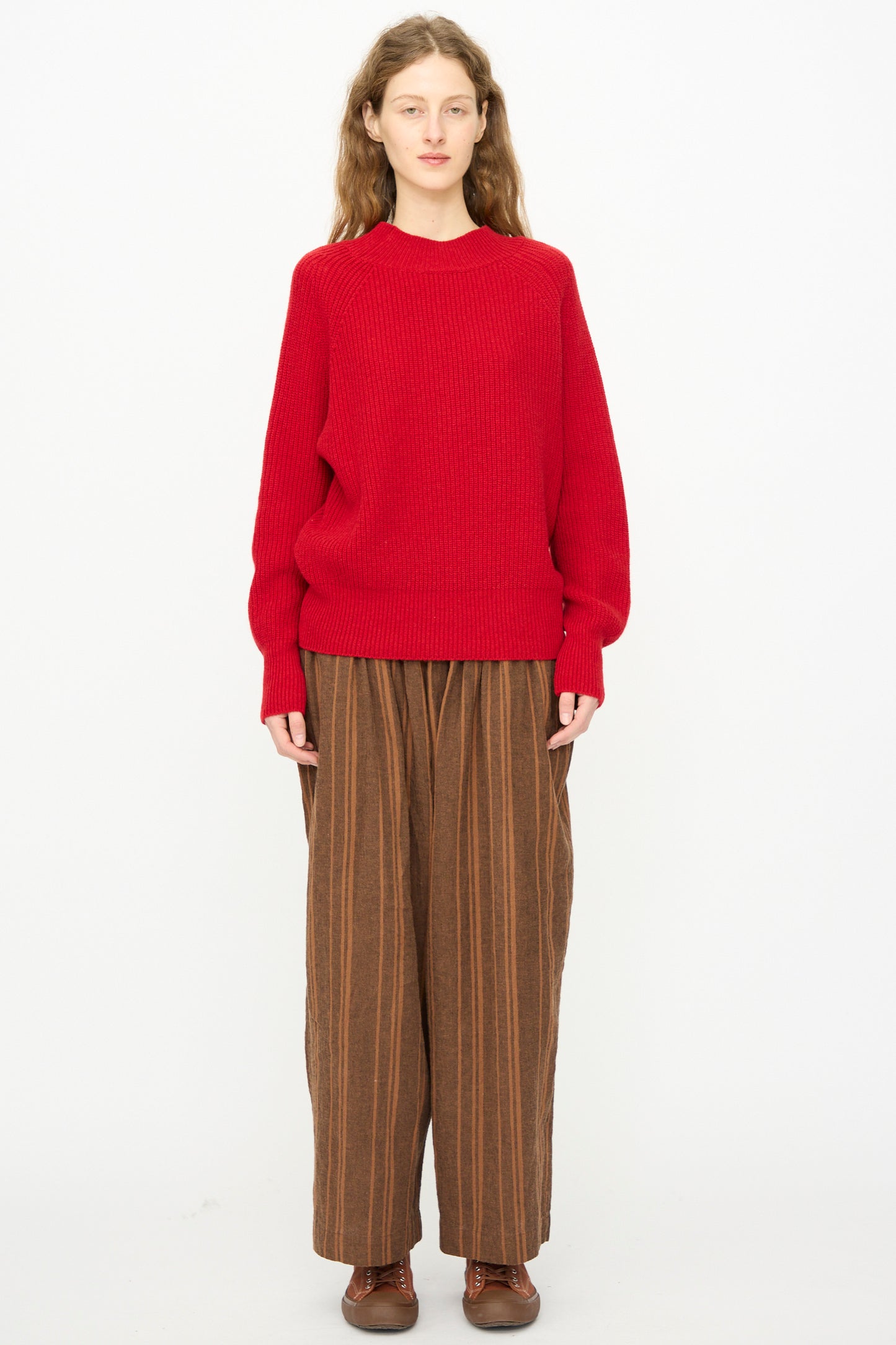 A person stands against a white background, wearing the Ichi Antiquités Wool Rib Knit Pullover in Red, wide brown striped pants, and brown shoes.