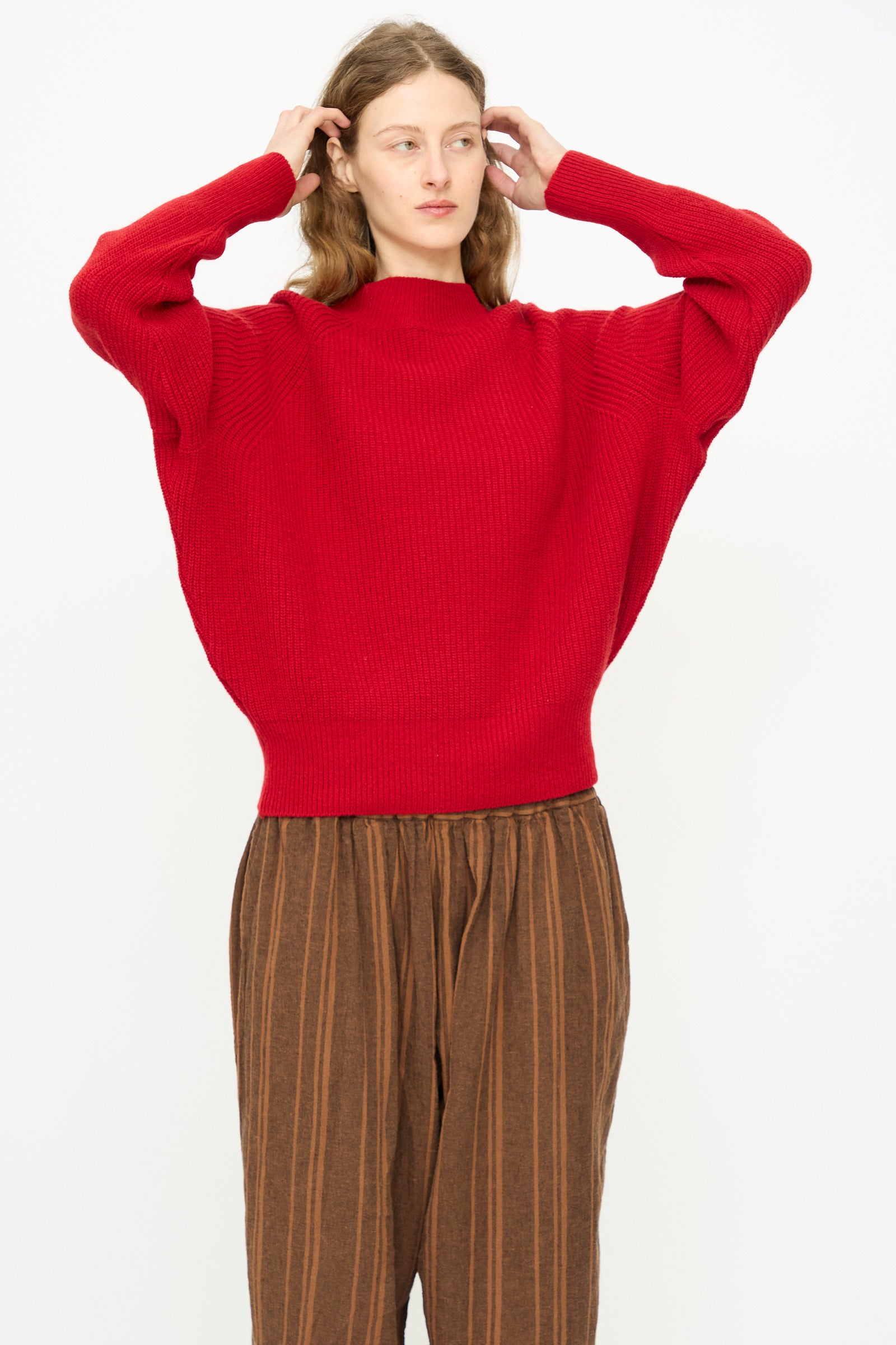 A person wears the Ichi Antiquités Wool Rib Knit Pullover in Red, paired with striped pants, standing against a plain white background and adjusting their hair.