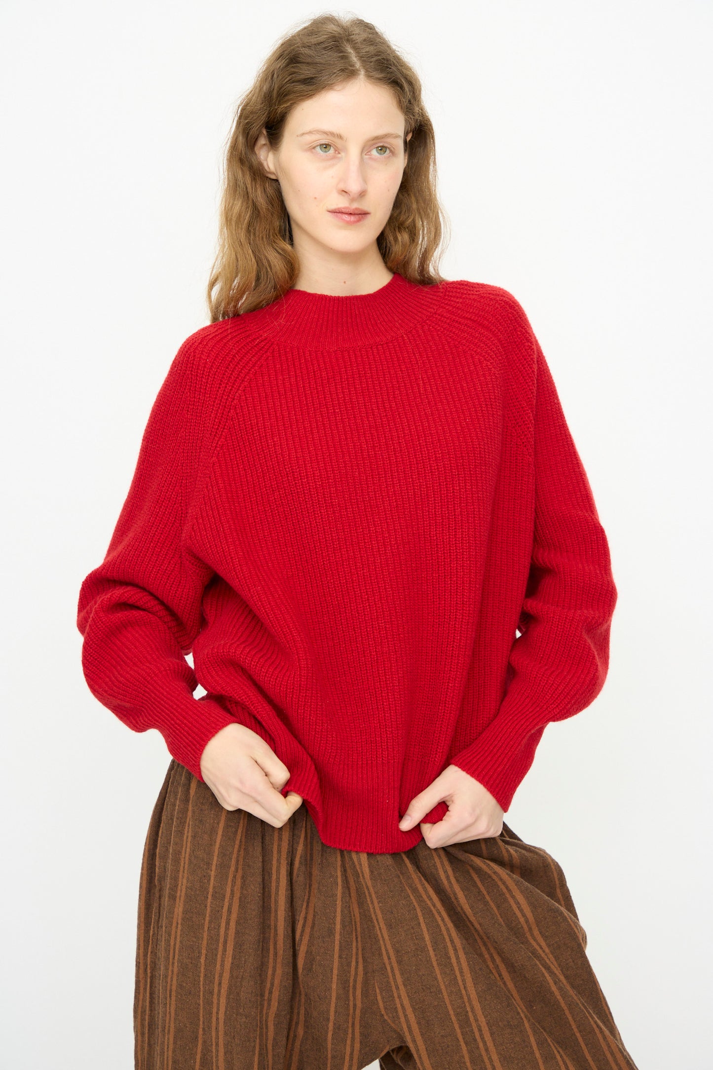 A person with long wavy hair wears the Ichi Antiquités Wool Rib Knit Pullover in Red and brown striped pants, standing against a plain white background.