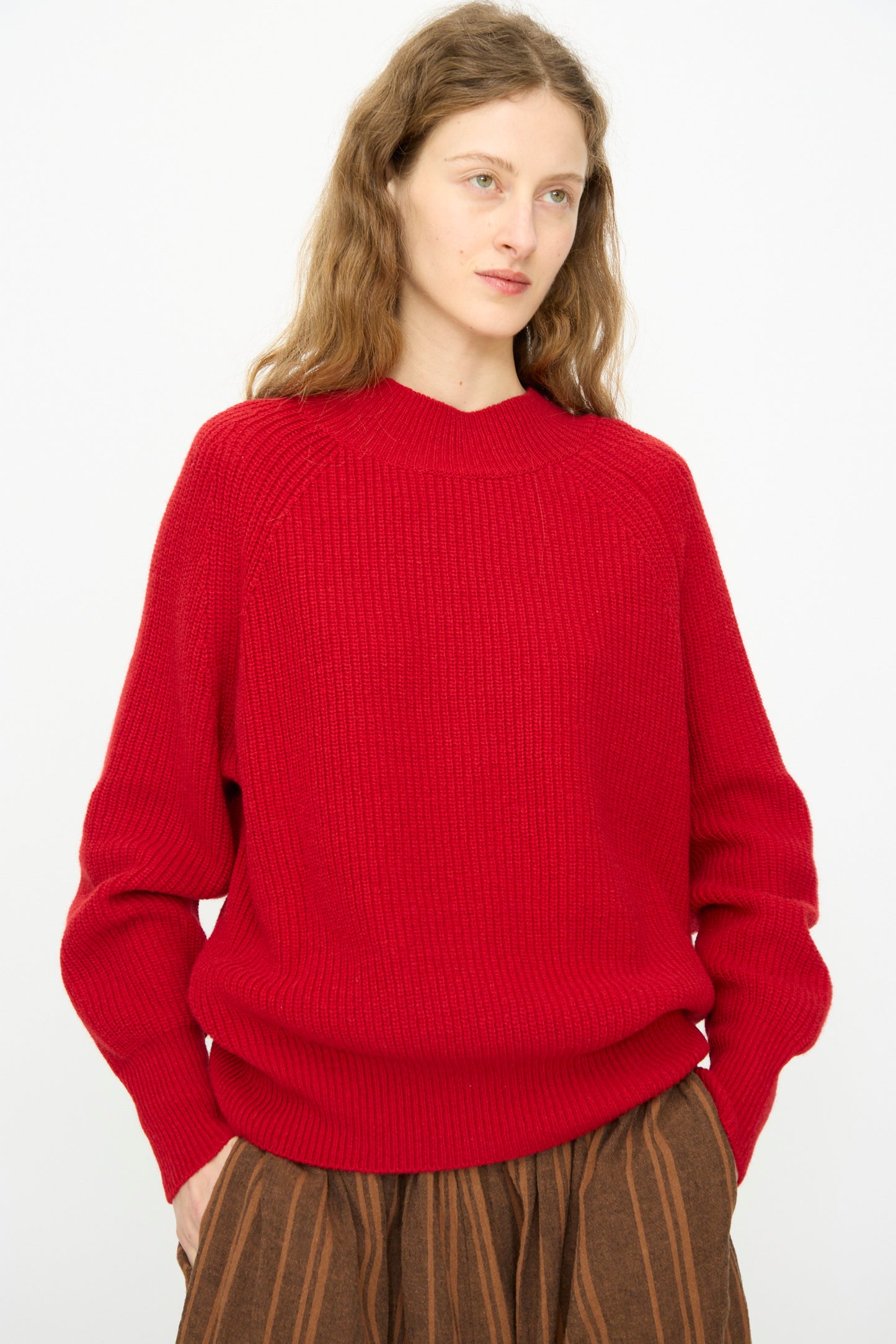 A person with long wavy hair wears the Wool Rib Knit Pullover in Red by Ichi Antiquités and a brown vertically striped skirt, standing against a plain light background.