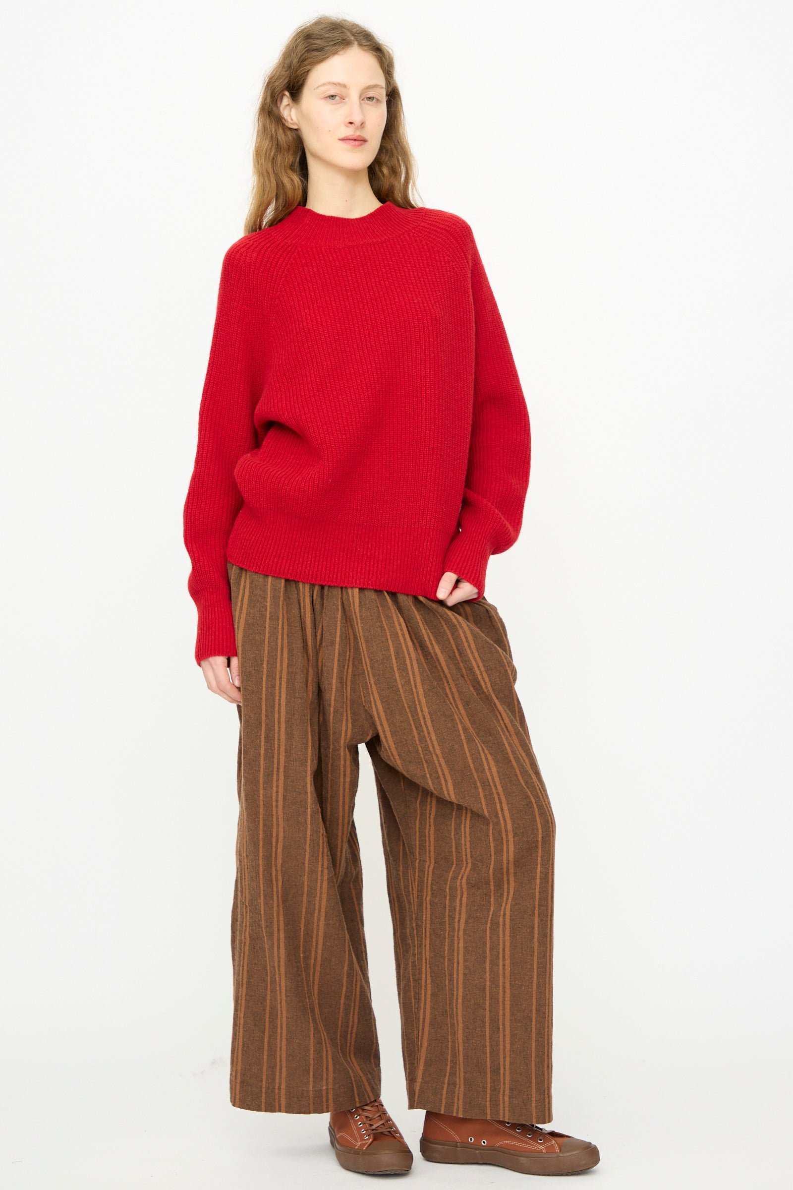 A person wears the Ichi Antiquités Wool Rib Knit Pullover in Red, wide brown striped pants, and brown shoes while standing against a plain white background.
