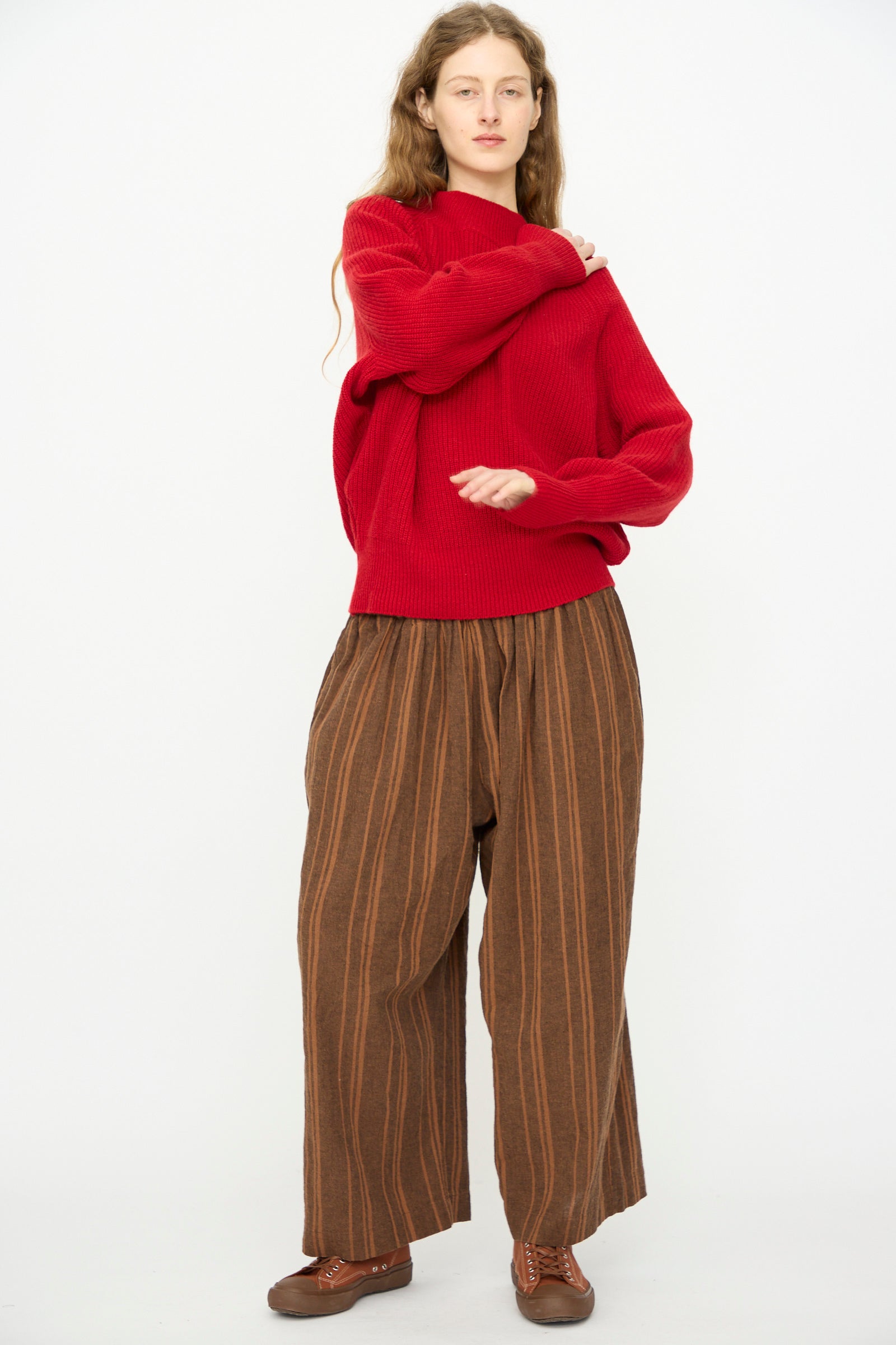 A person stands against a white background wearing the Ichi Antiquités Wool Rib Knit Pullover in Red, paired with loose brown striped pants and brown shoes, with one arm crossed over their chest.