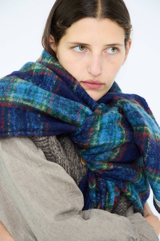 A person with long hair wears the Ichi Antiquités Wool Tartan Shaggy Stole in Navy, featuring shades of blue, green, and red, draped over a textured beige and gray outfit.