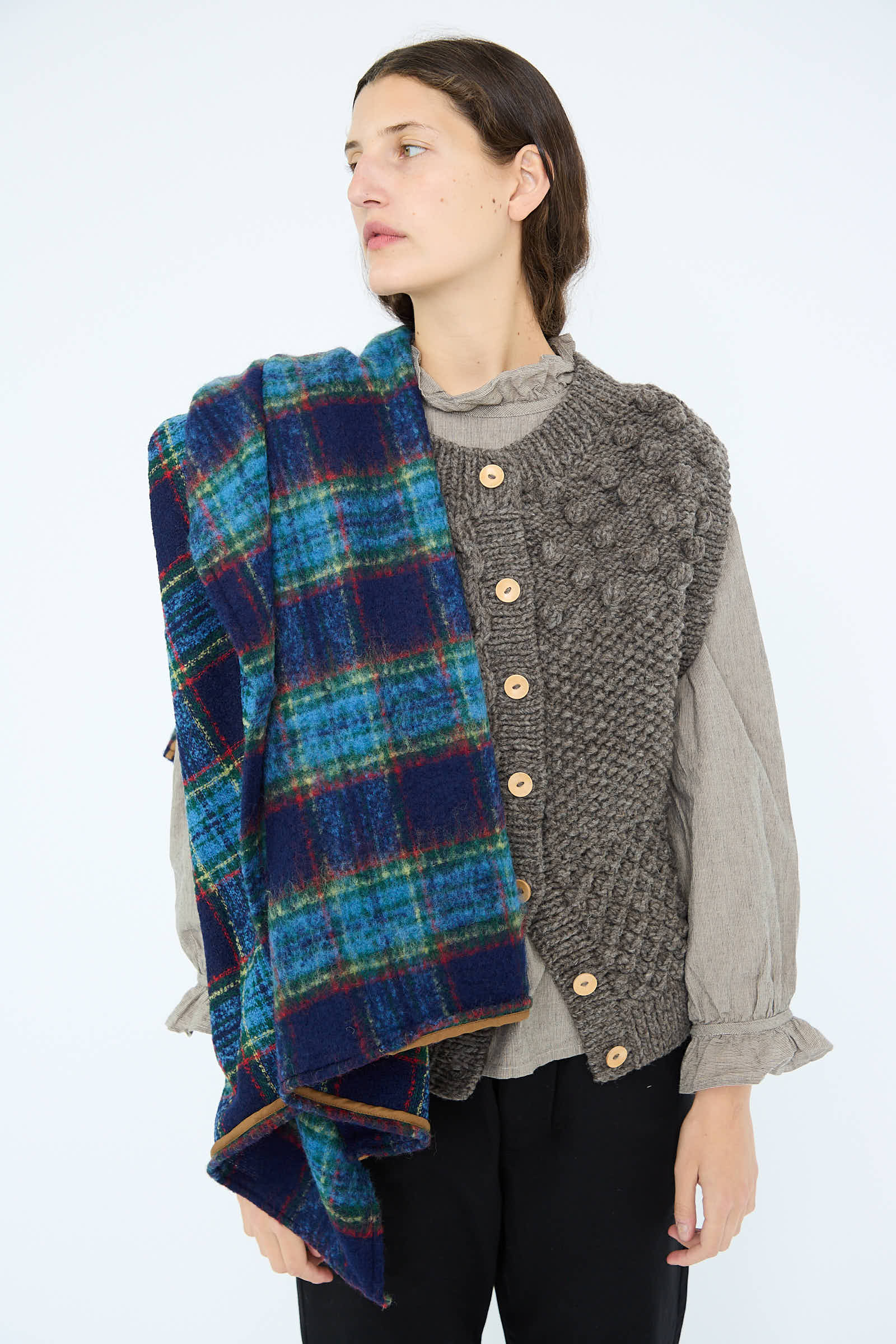 A person wearing a textured vest and draping the Wool Tartan Shaggy Stole in Navy by Ichi Antiquités, styled as a blue plaid blanket, over their shoulder, gazing to the side against a plain backdrop.