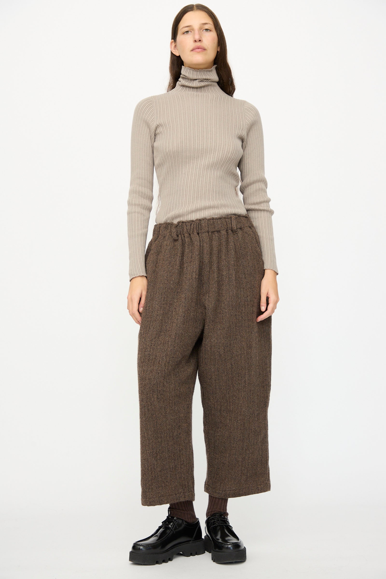 A person stands against a plain white background wearing Wool Herringbone Pants in Brown by Ichi Antiquités, paired with a beige ribbed turtleneck sweater, dark socks, and black shoes.