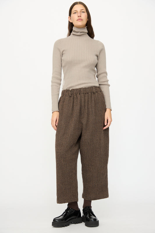 A person stands against a plain white background wearing Wool Herringbone Pants in Brown by Ichi Antiquités, paired with a beige ribbed turtleneck sweater, dark socks, and black shoes.