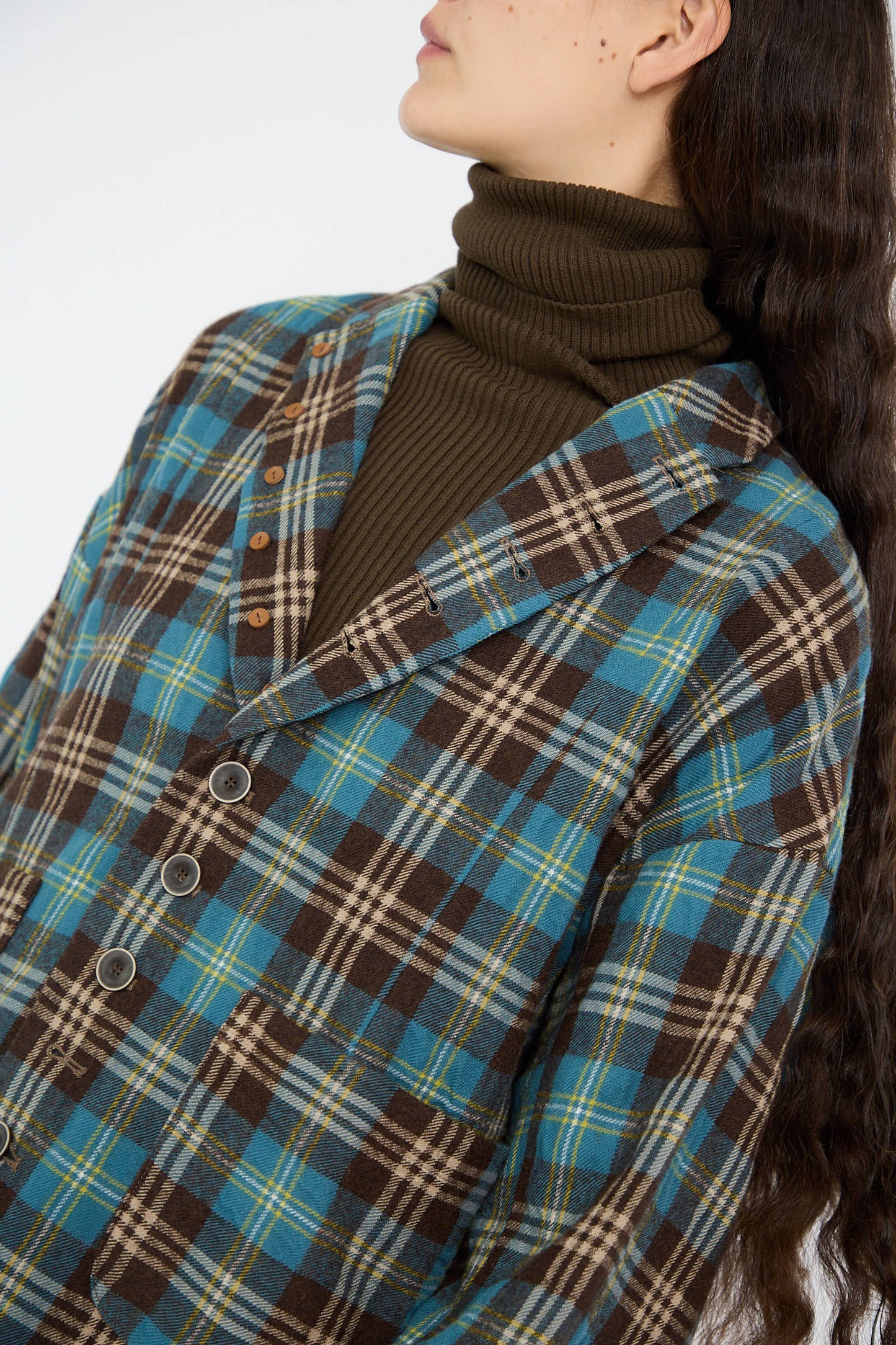 A person wearing the Ichi Antiquités Wool Washer Tartan Jacket in Blue, featuring a notched collar and turtleneck, with long hair flowing, depicted from the shoulders up.