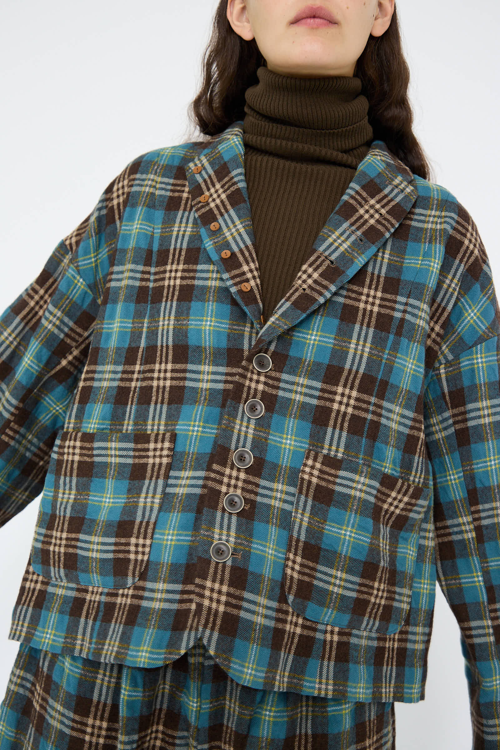 Dressed in the Wool Washer Tartan Jacket in Blue by Ichi Antiquités, a person pairs it effortlessly with a washed wool turtleneck featuring large buttons and a notched collar.