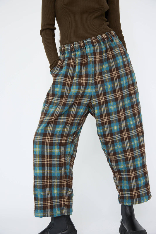 A person wearing the Wool Washer Tartan Pant in Blue by Ichi Antiquités, featuring an elasticated waist, paired with a dark long-sleeve shirt and black boots, stands against a white background.