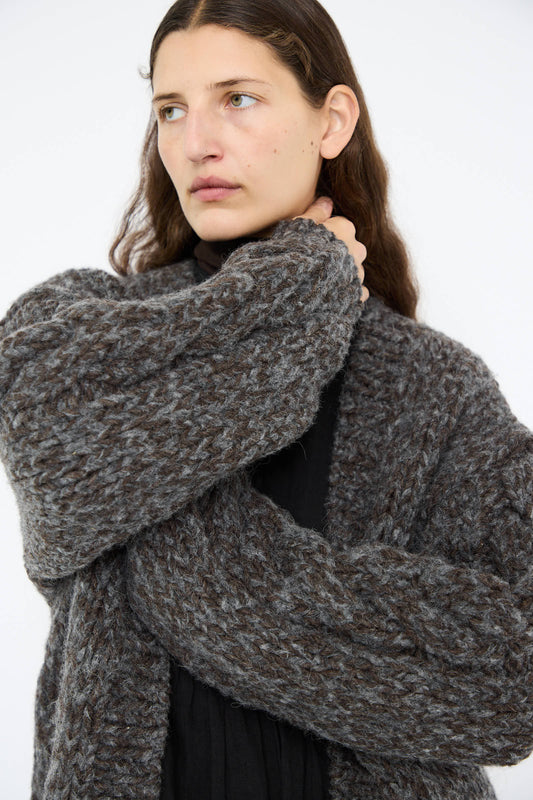 Dressed in an Ichi Antiquités Wool and Alpaca Hand Knit Cardigan in Charcoal over a black top, a person gazes to the side with one hand near their neck.