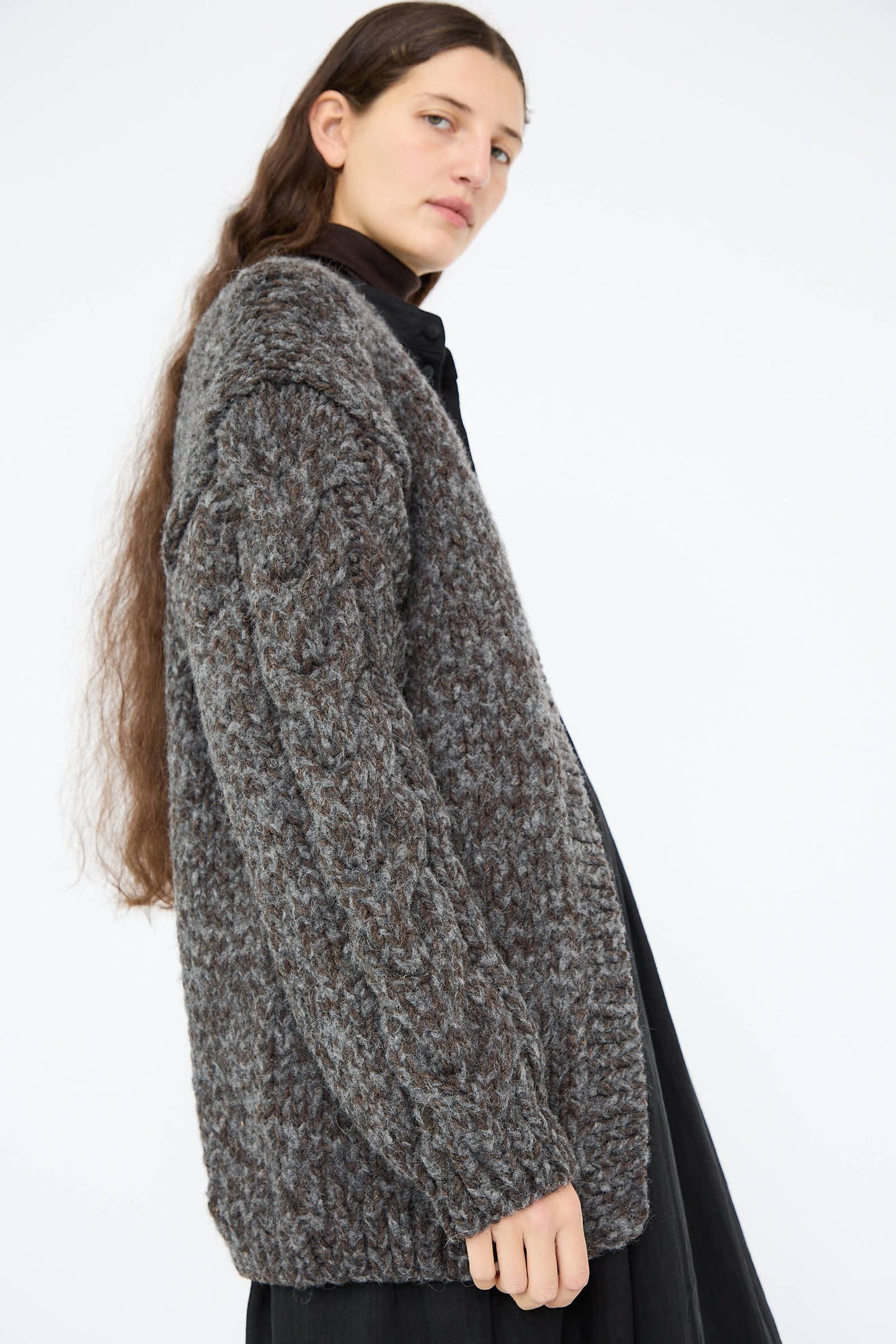 A person with long hair wearing a thick, textured wool and alpaca hand-knit cardigan in charcoal by Ichi Antiquités, paired with a black outfit, looking to the side.