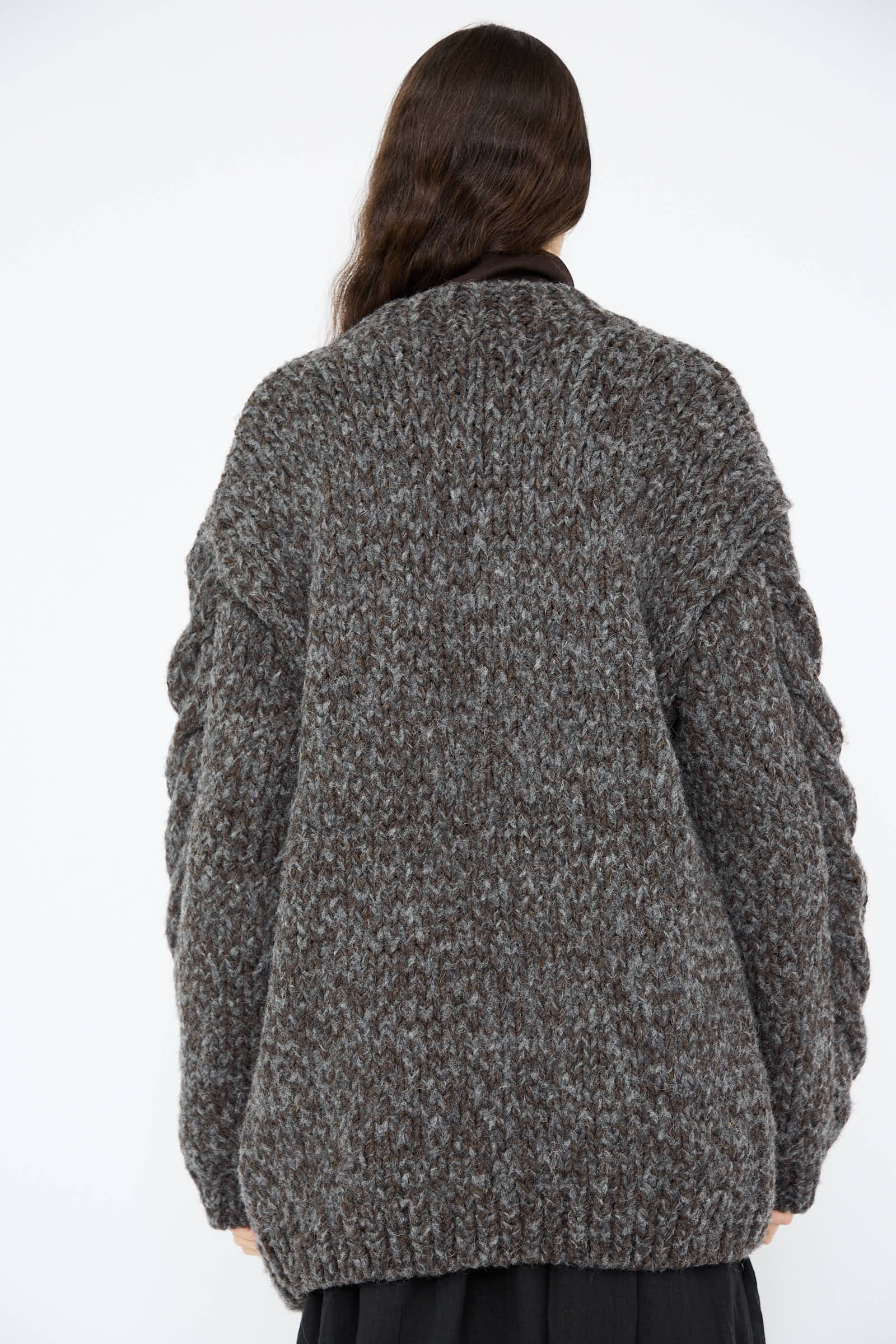 A person with long dark hair is shown from the back, wrapped in the Wool and Alpaca Hand Knit Cardigan in Charcoal by Ichi Antiquités, featuring intricate cable knit detailing.