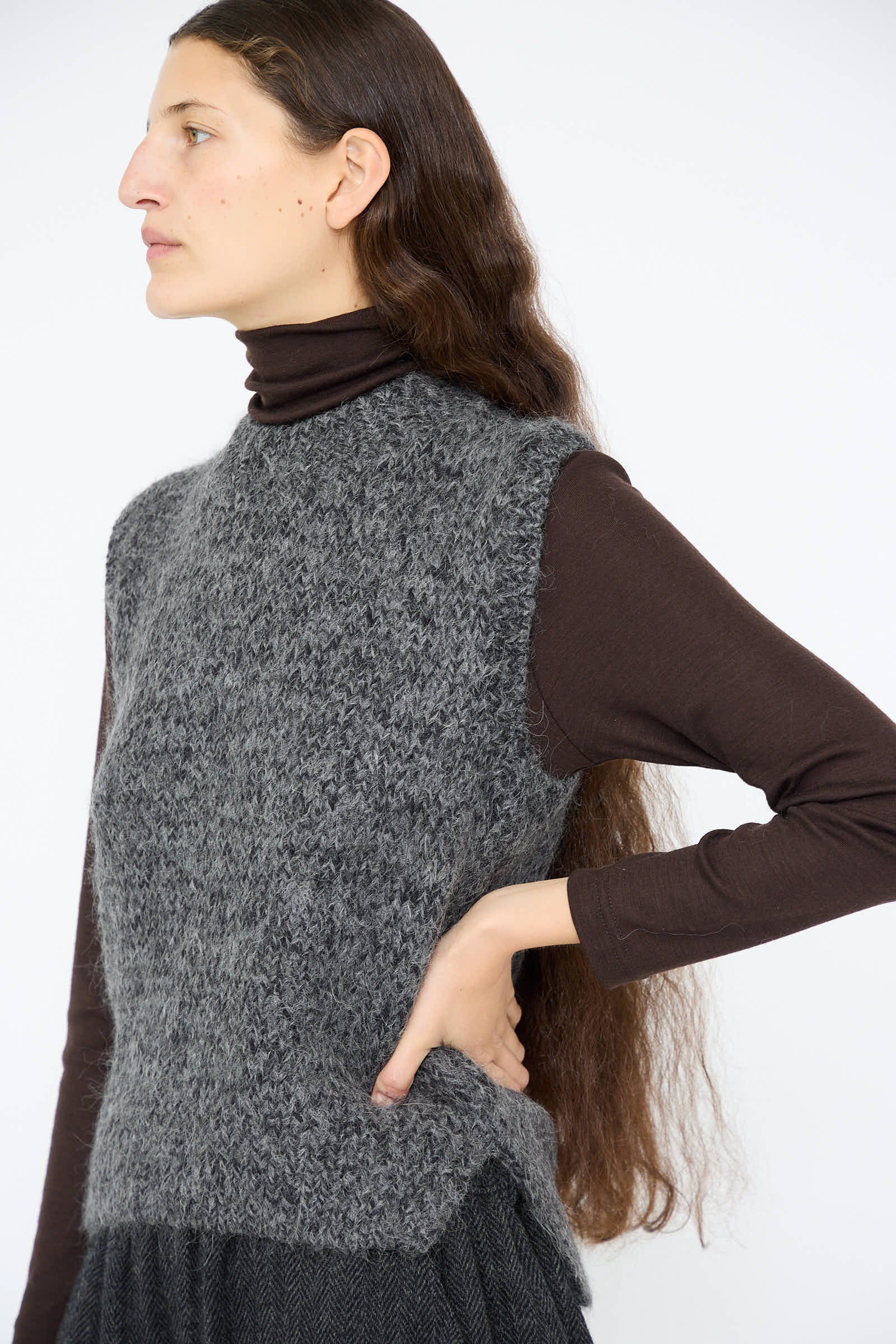 An individual with long hair wears a Hand Knit Wool and Alpaca Vest in Gray by Ichi Antiquités, layered over a dark long-sleeve turtleneck, standing against a plain background.