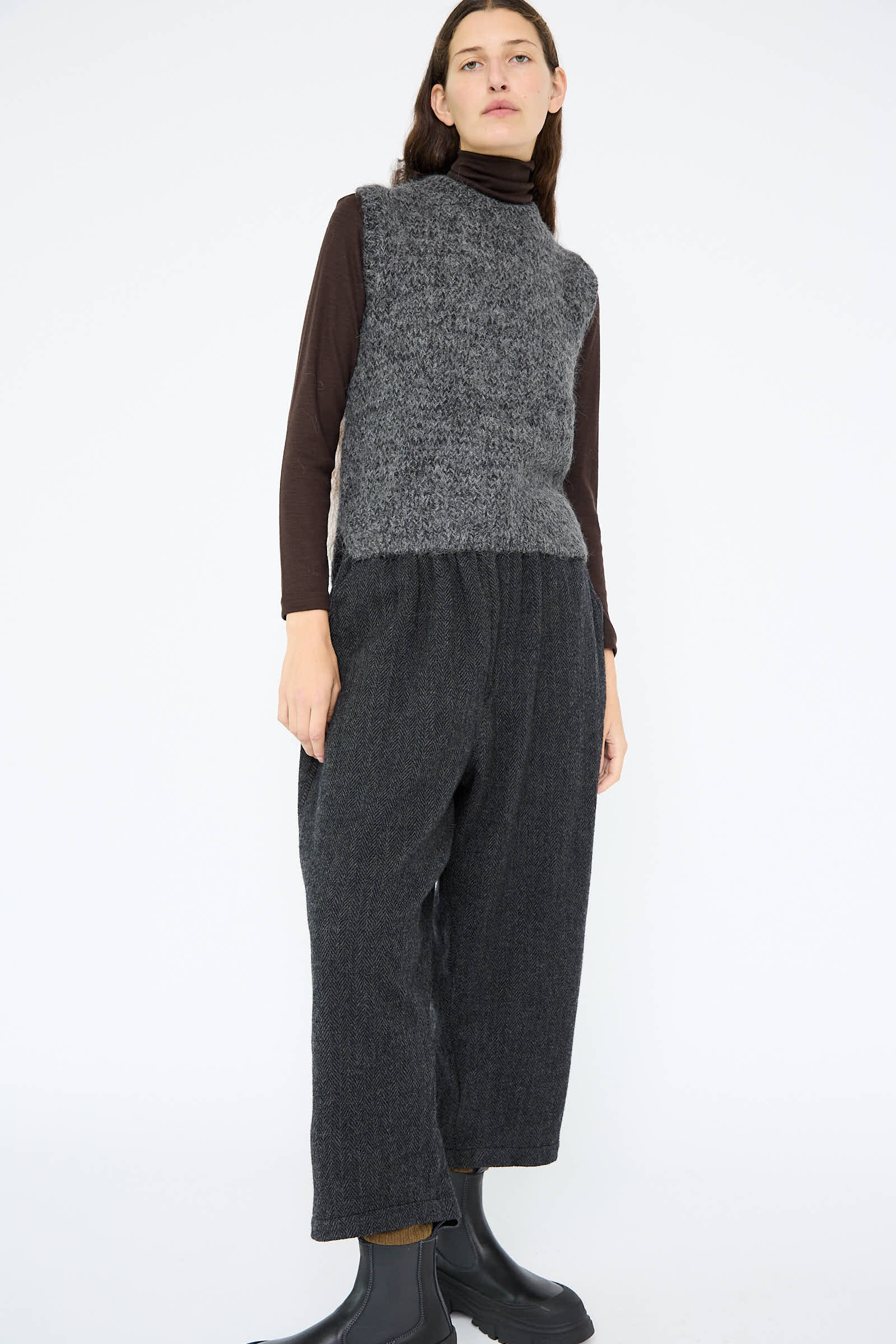 A person wearing the Hand Knit Wool and Alpaca Vest in Gray by Ichi Antiquités, layered over a dark long-sleeve top and paired with loose gray pants for a relaxed fit, stands against a plain white background.