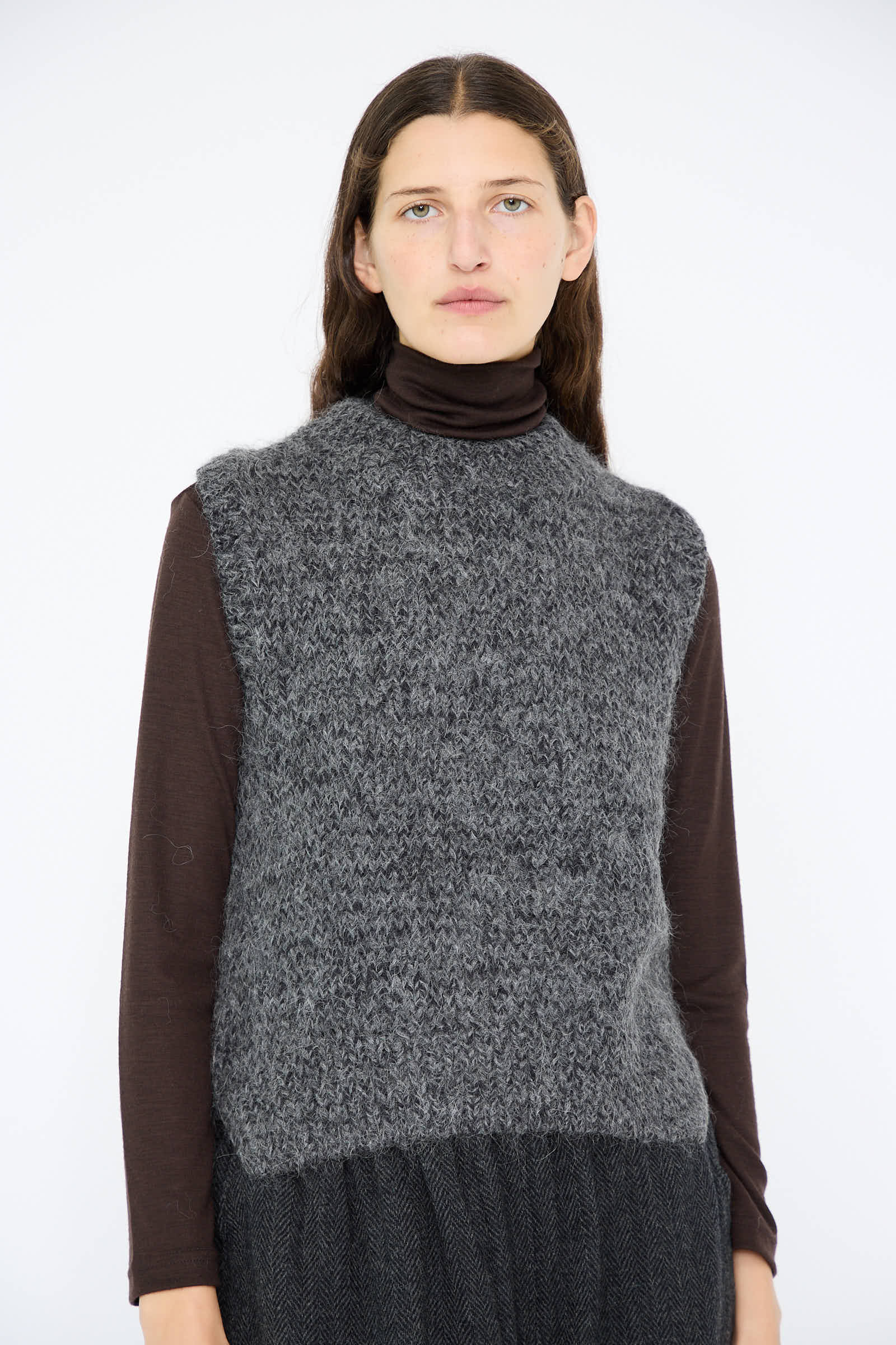 A person with long dark hair models against a plain white background, wearing the Hand Knit Wool and Alpaca Vest in Gray by Ichi Antiquités over a dark turtleneck. The vest's relaxed fit introduces an effortlessly casual elegance to their ensemble.