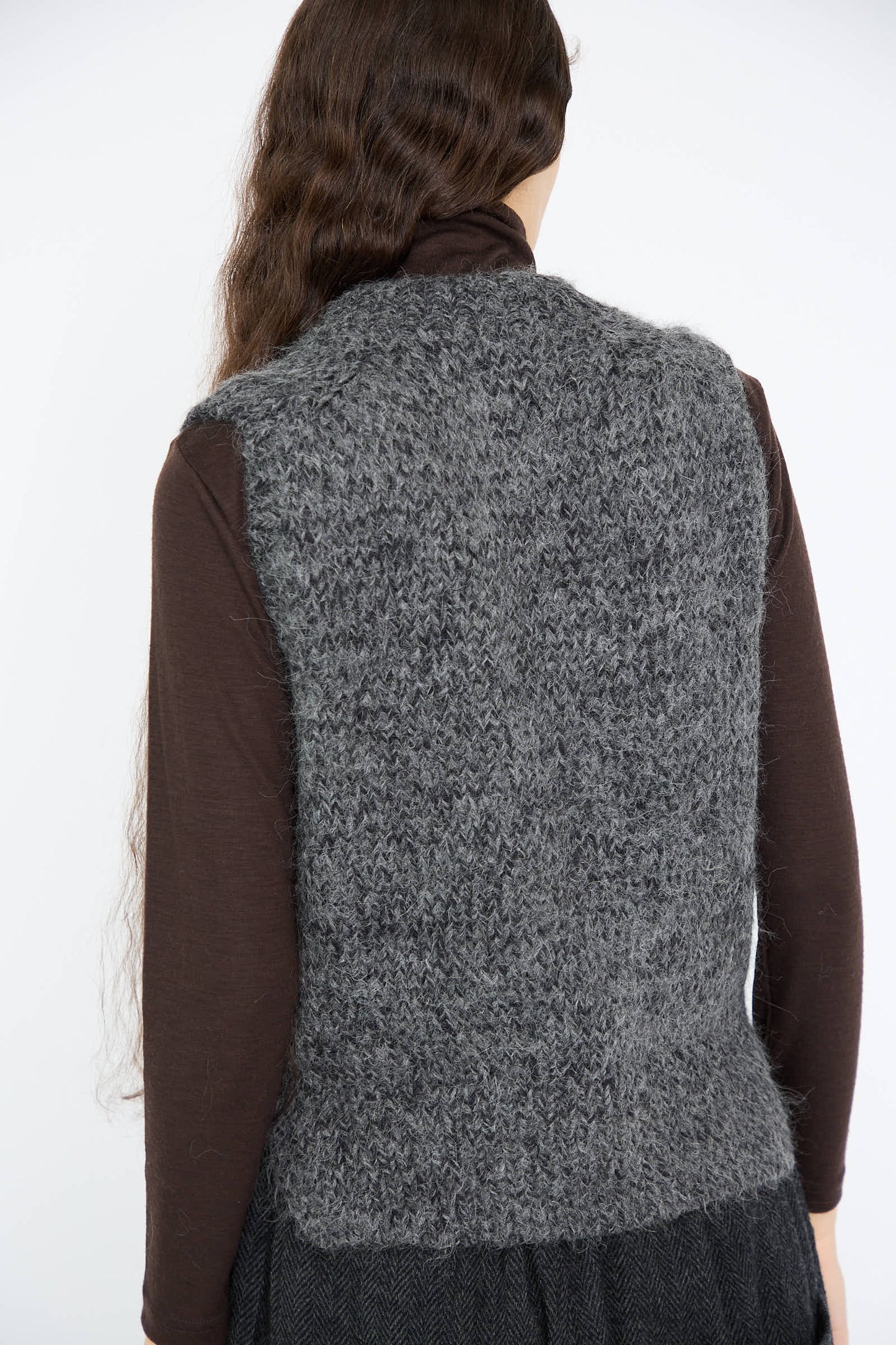 A person with long brown hair, wearing the Ichi Antiquités Hand Knit Wool and Alpaca Vest in Gray over a dark long-sleeve shirt, is facing away.