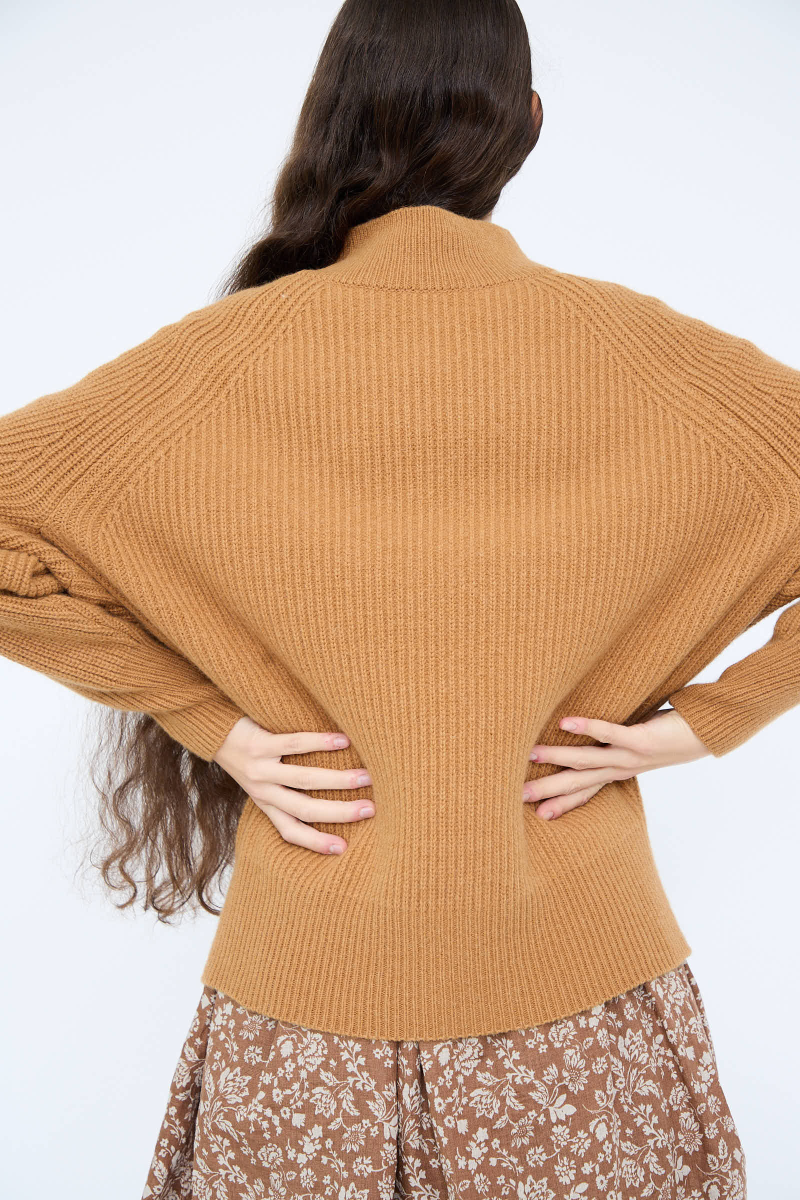 A person with long brown hair is wearing the Wool and Angora Ridge Knitted Pullover in Camel by Ichi Antiquités, paired with a floral skirt. They have their back turned and hands on hips.