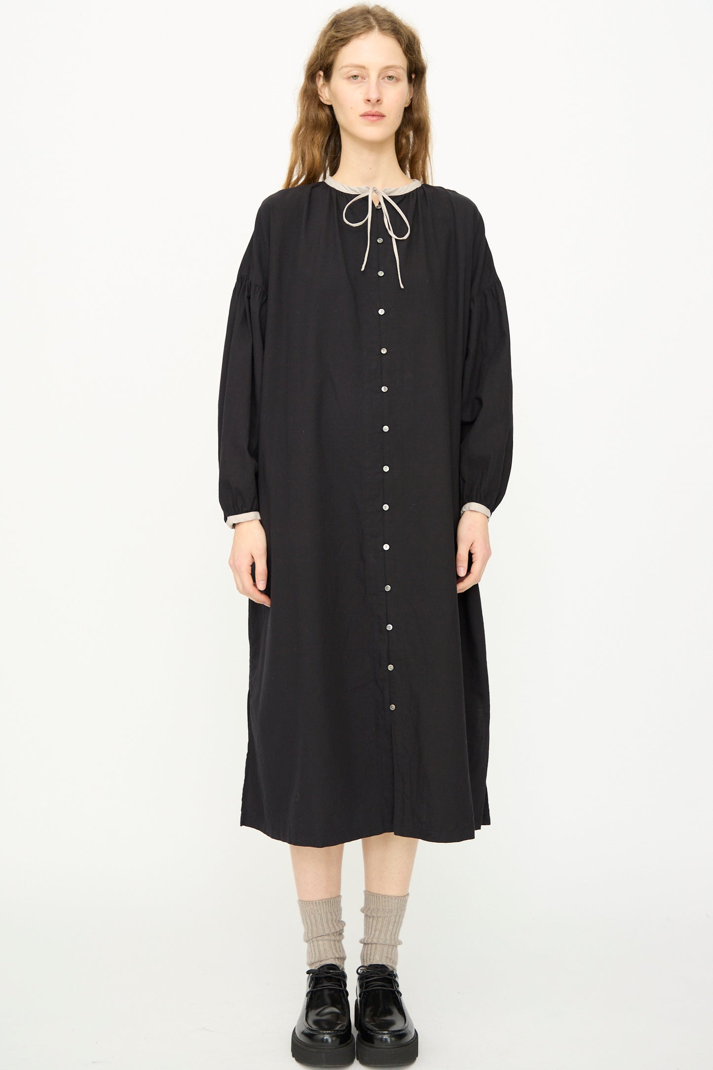 A person stands facing forward against a plain white background, wearing the Ichi Antiquités Cotton Cashmere Viyella Dress in Black with a neck tie, paired with gray socks and black shoes.