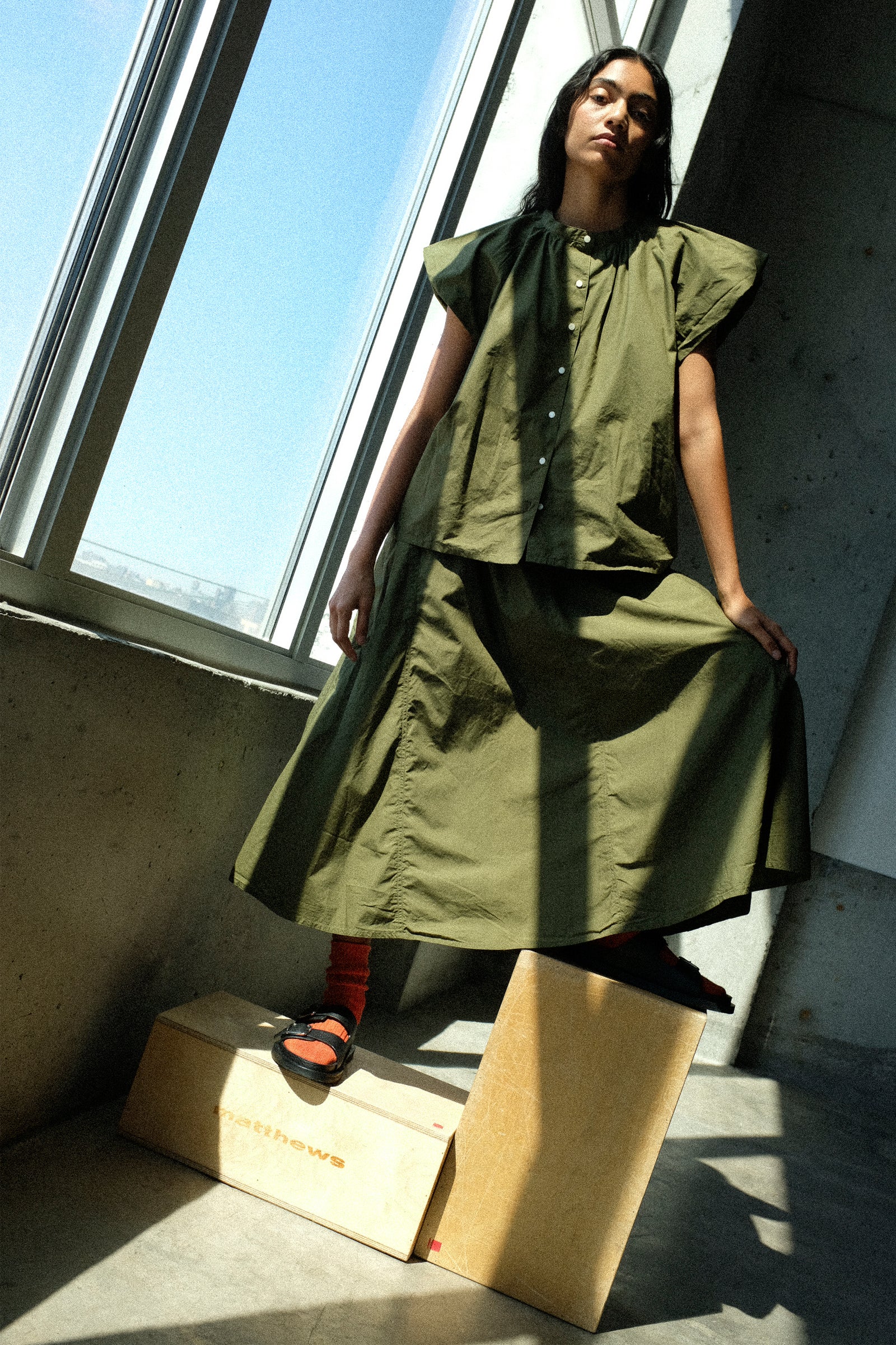 Woman wearing Ichi Antiquités Cotton Typewriter Shirt in Khaki, and Cotton Typewriter Skirt in Khaki standing on apple boxes in a room with large windows and concrete walls.