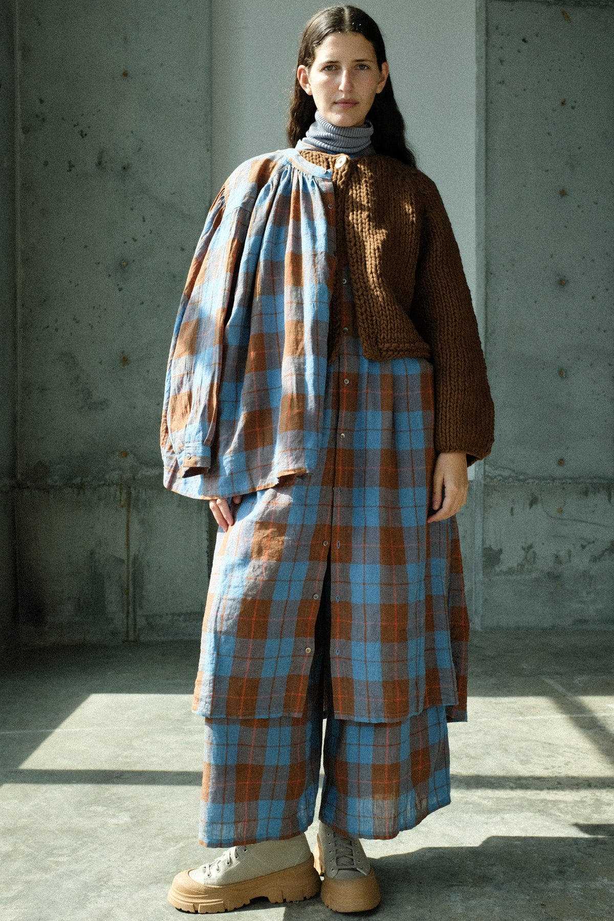 A woman in a sun filled studio wearing Ichi Antiquites Linen Cotton Check Dress in Blue, Cotton Rib Turtleneck in Light Gray, Wool Hand Knit Pullover in Brown, and Linen Cotton Check Pants in Blue.