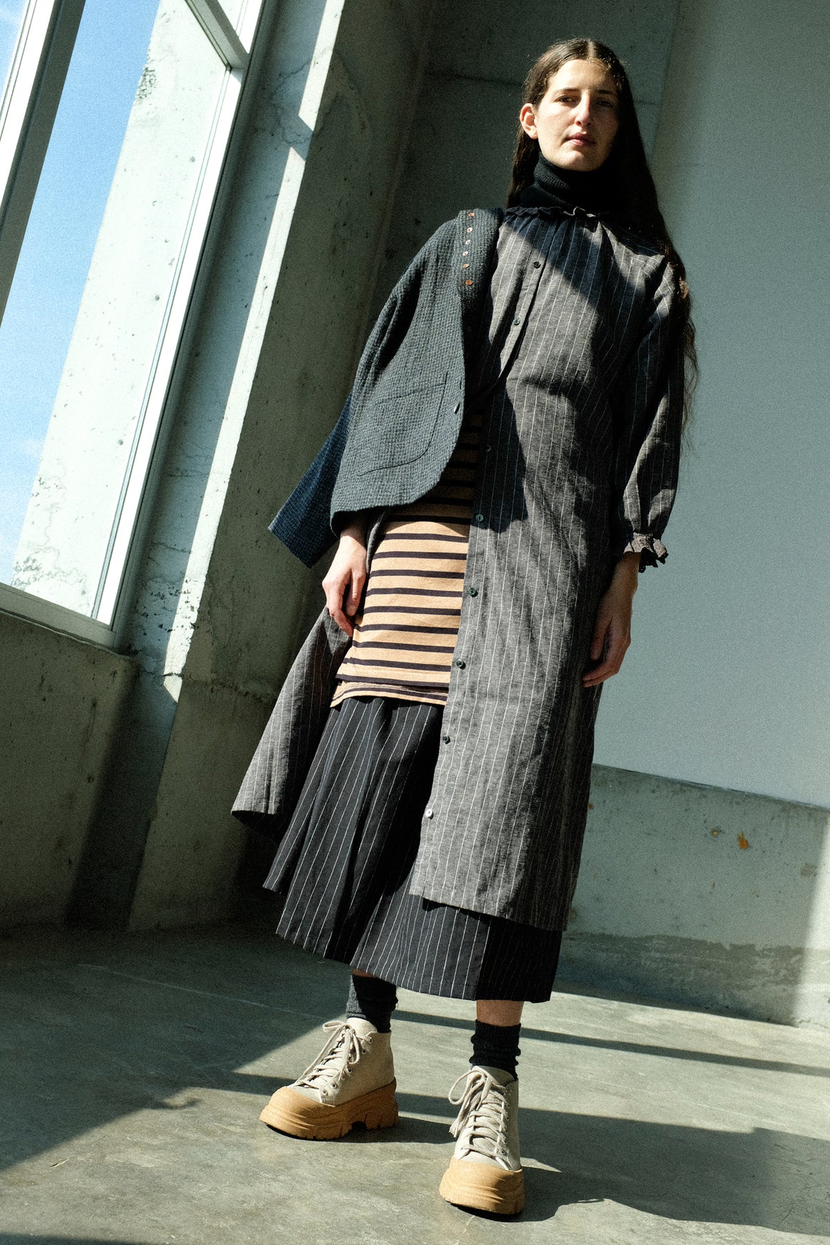 A woman standing in front of a window wearing Ichi Antiquités Wool Gauze Houndstooth Jacket in Charcoal over the Pinstripe Dress in Charcoal, with the Cotton Stripe Pullover in Mocha and Navy tied around her waist.