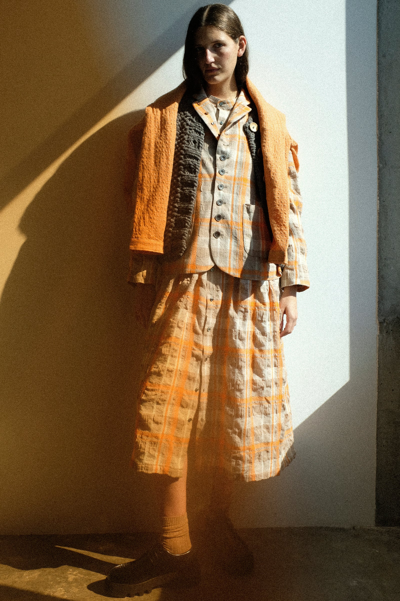 A woman wearing orange tartan from the FW25 Ichi Antiquités collection against a white wall and an orange filter on the side.