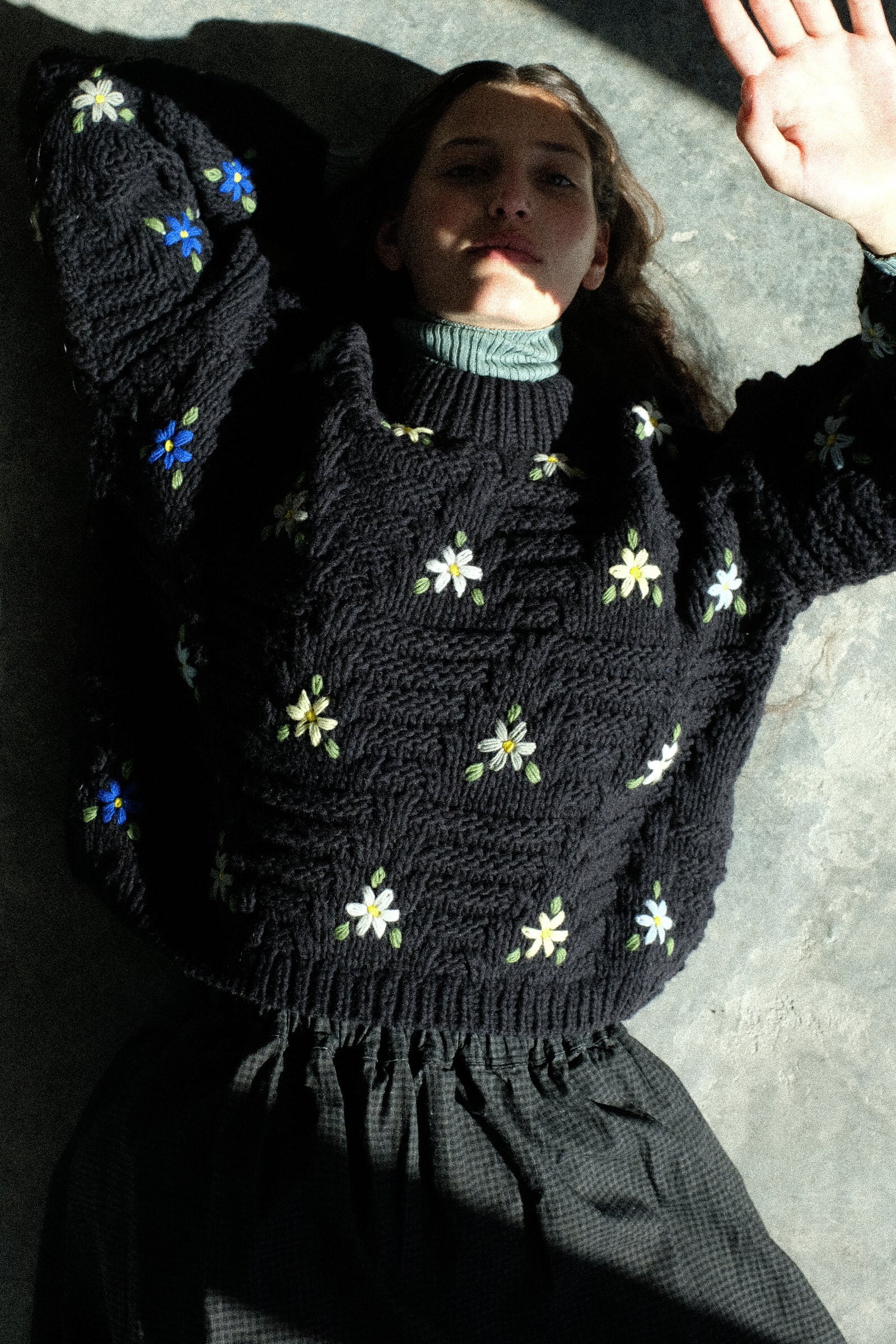A woman laying on the ground wearing Wool Hand-knit Pullover with Flowers in Black, holding her hand up to block the sun.