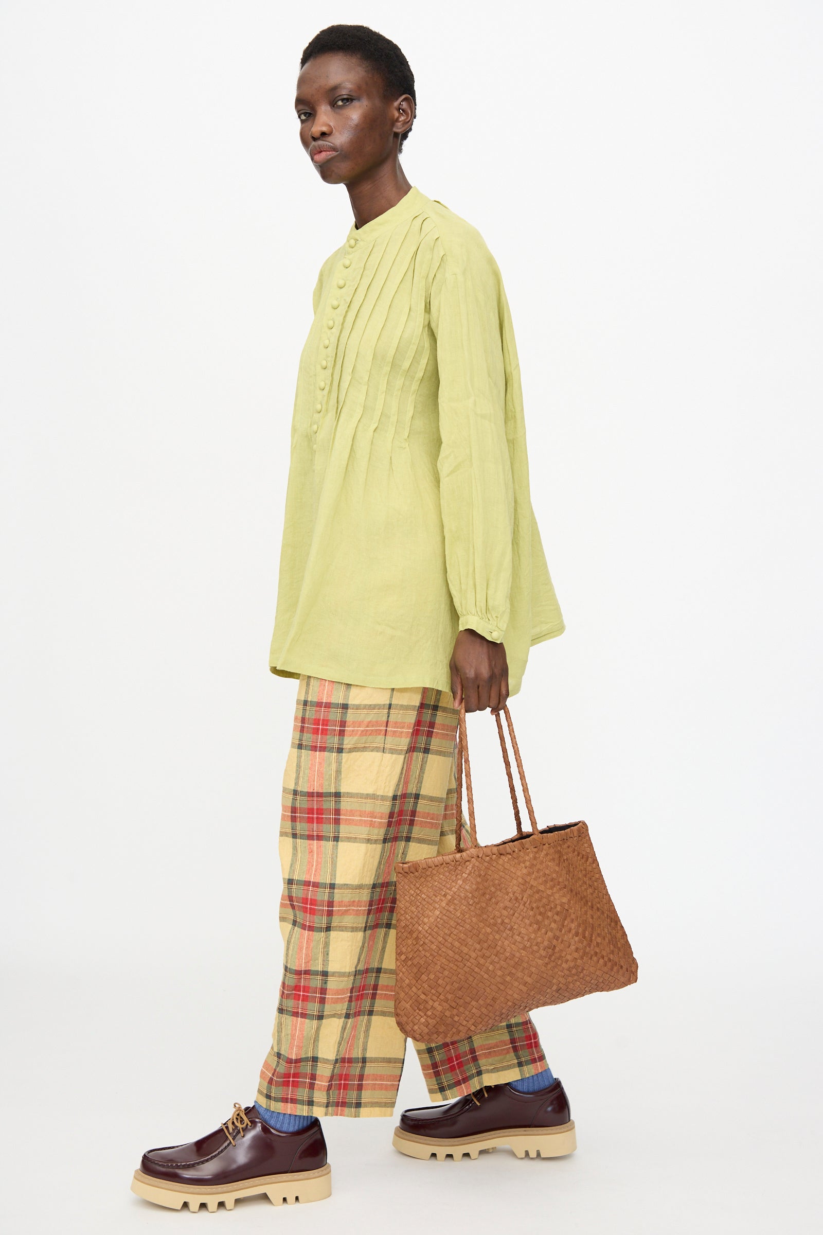 A person wears a light green long-sleeve blouse, plaid pants, platform shoes, and carries the Ichi Antiquités Leather Tote Bag in Brown against a plain white background.