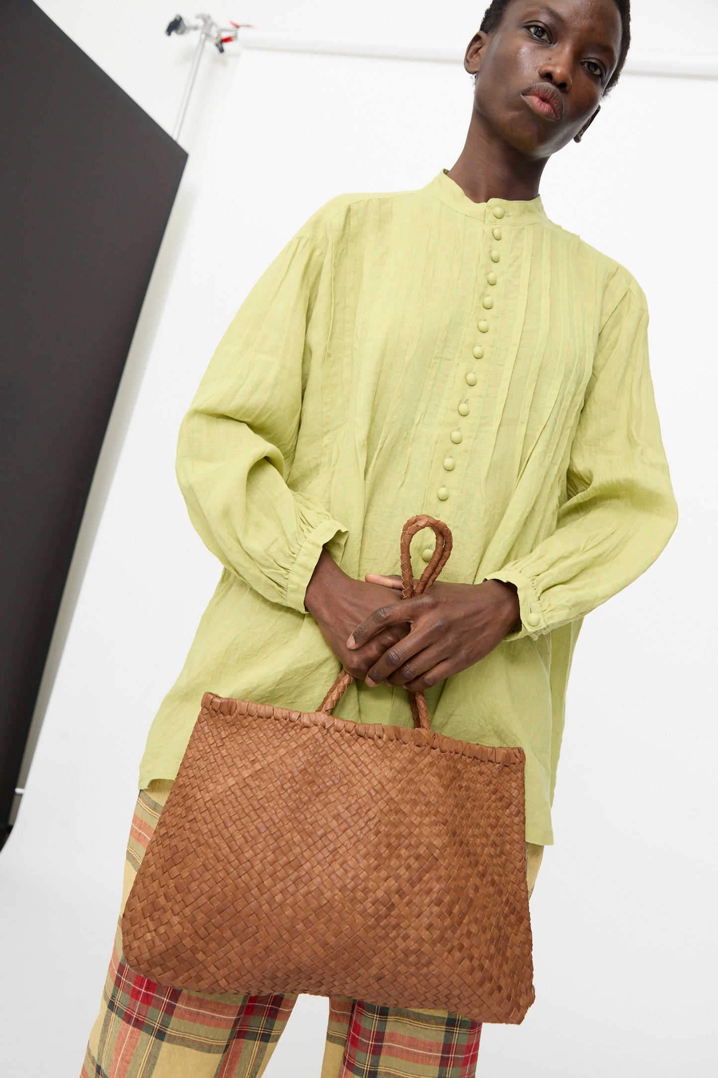 In a studio setting, someone holds the Ichi Antiquités Leather Tote Bag in Brown while wearing a light green button-up blouse and plaid pants.