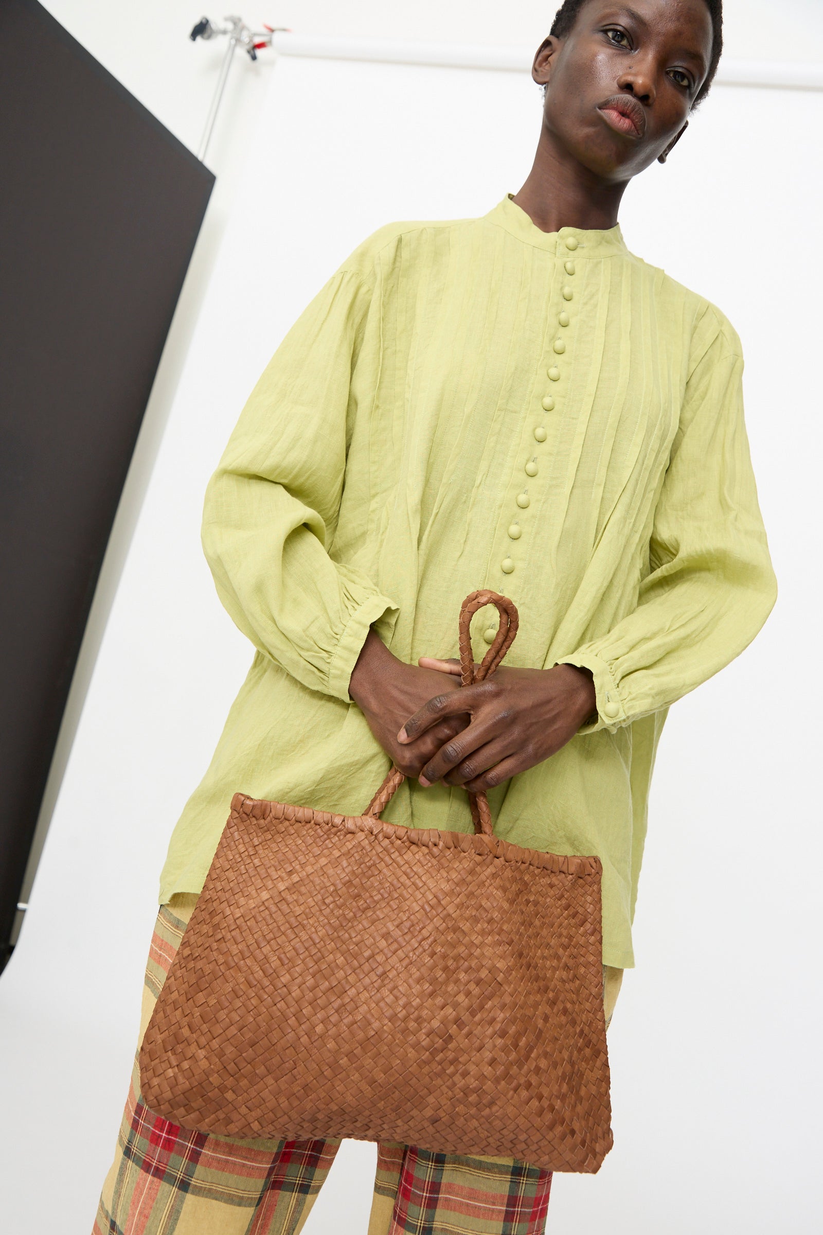 In a studio setting, someone holds the Ichi Antiquités Leather Tote Bag in Brown while wearing a light green button-up blouse and plaid pants.
