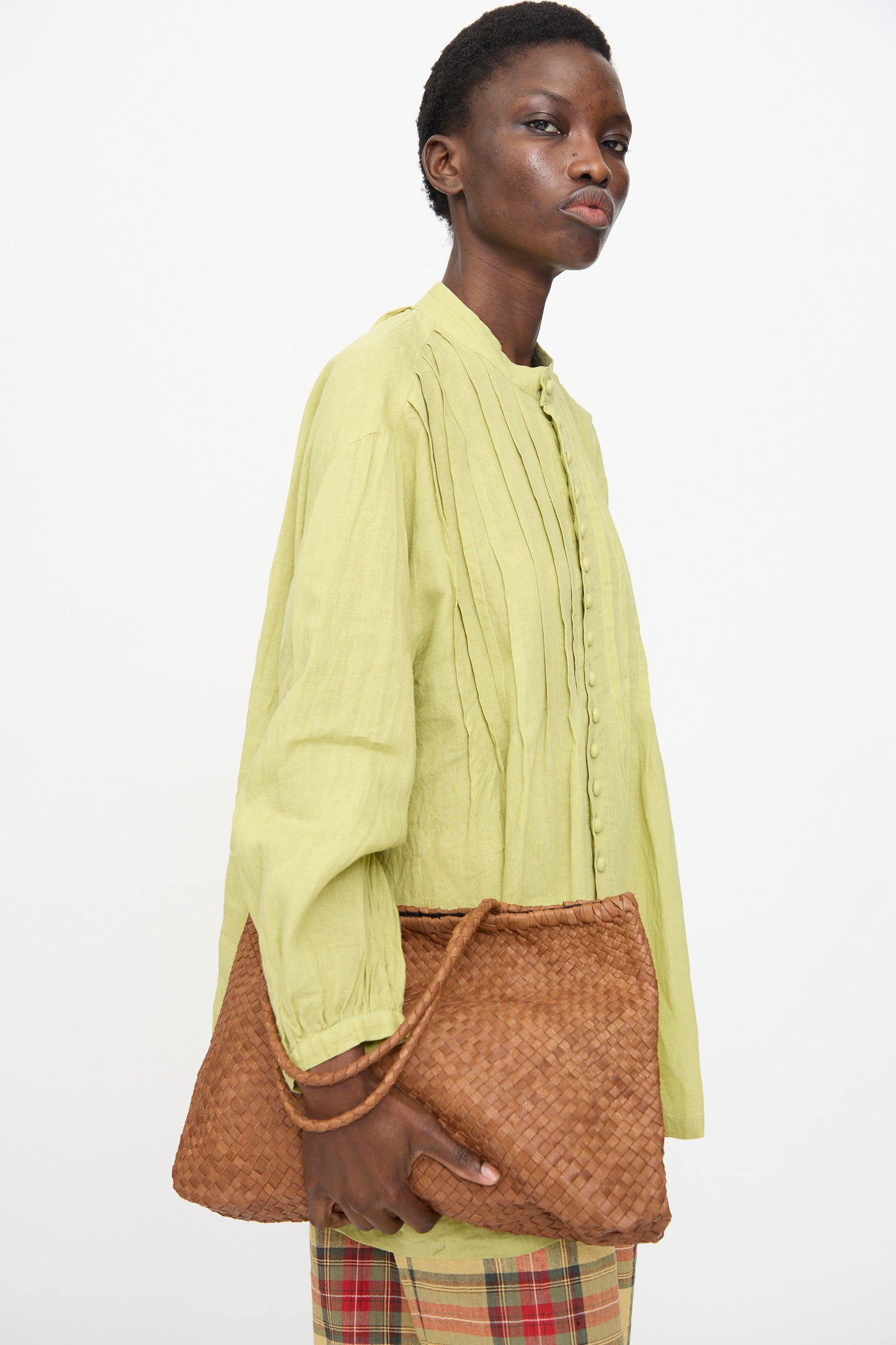 A person holds the Ichi Antiquités Leather Tote Bag in Brown while wearing a light green pleated blouse and plaid pants against a plain white background.