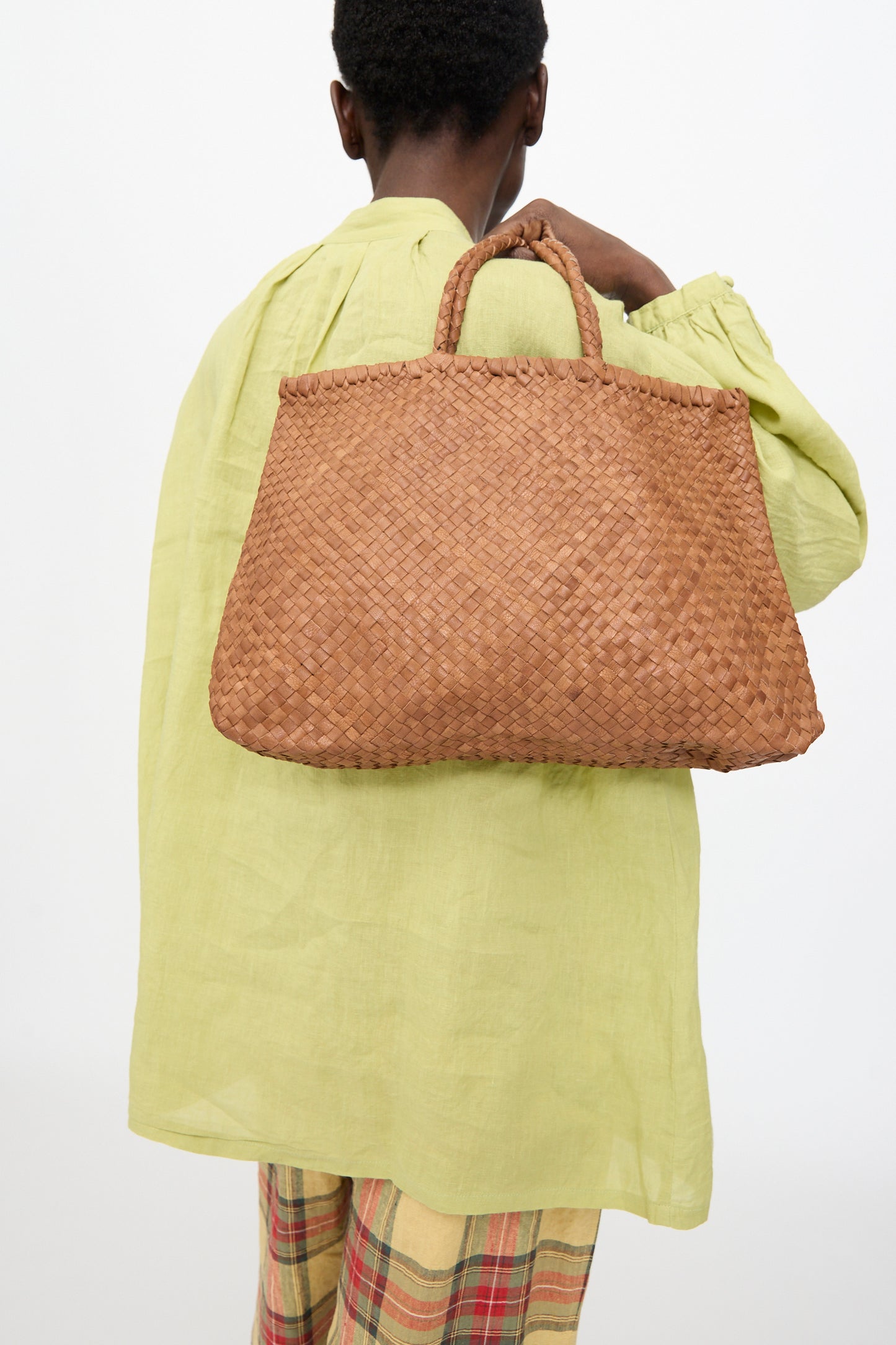 A person with short hair, dressed in a light green top and plaid pants, carries the Ichi Antiquités Leather Tote Bag in Brown over their shoulder, facing away from the camera.