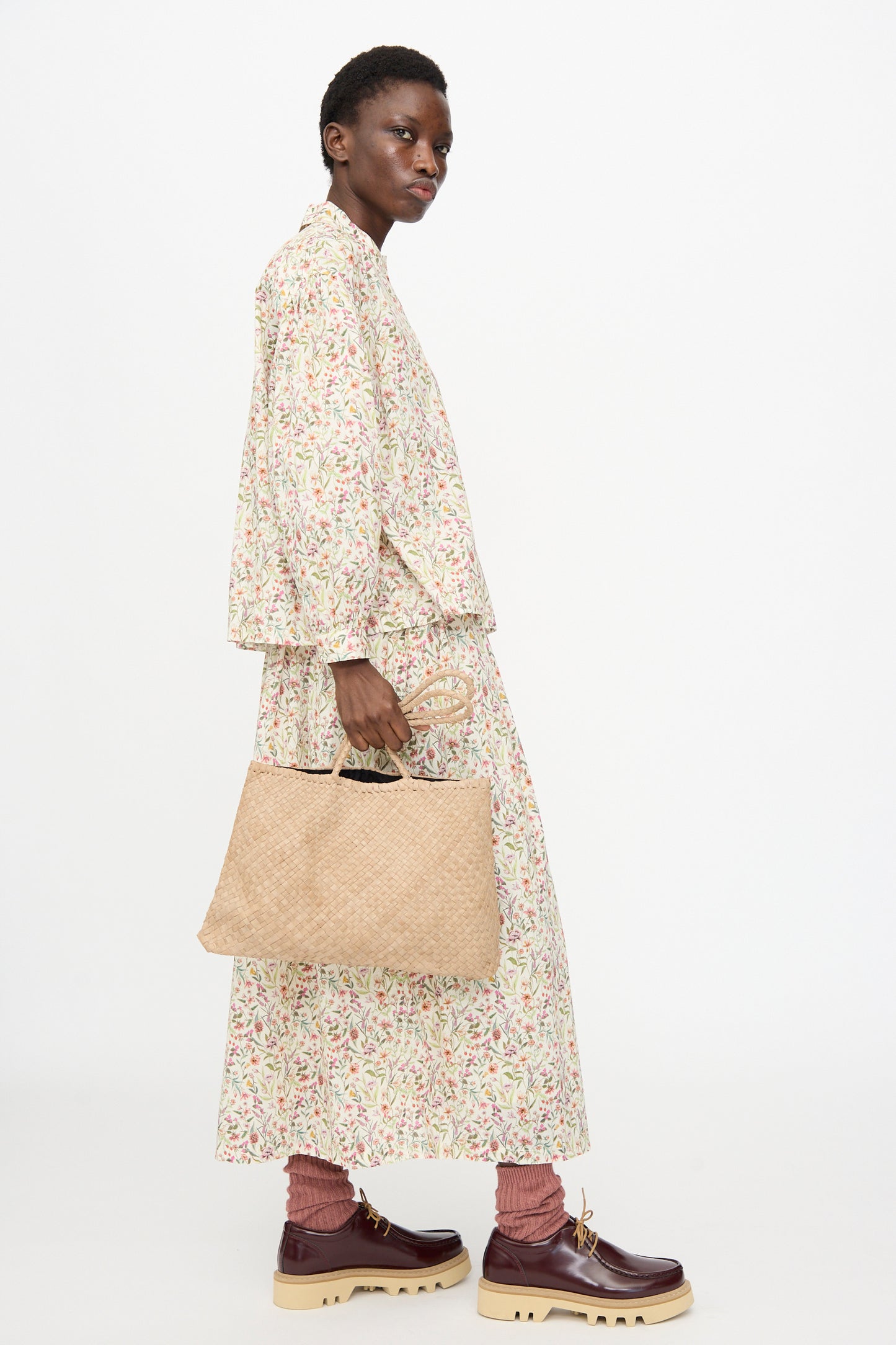 Person in profile wears a long floral dress, chunky brown shoes, pink socks, and carries the Ichi Antiquités Leather Tote Bag in Greige against a plain white background.