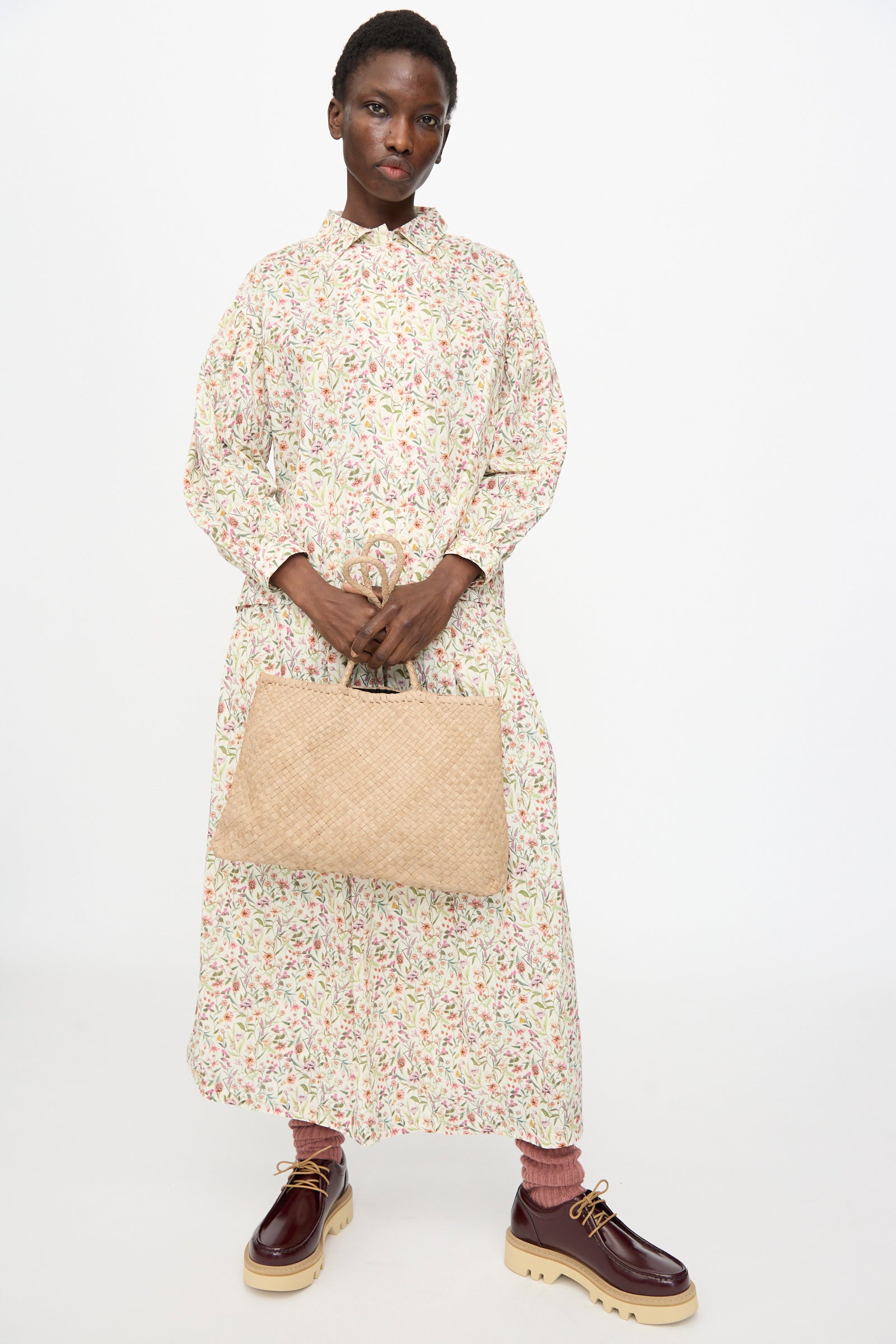 Person standing against a plain background, wearing a long floral dress, pink pants, burgundy platform shoes, and holding the Ichi Antiquités Leather Tote Bag in Greige.