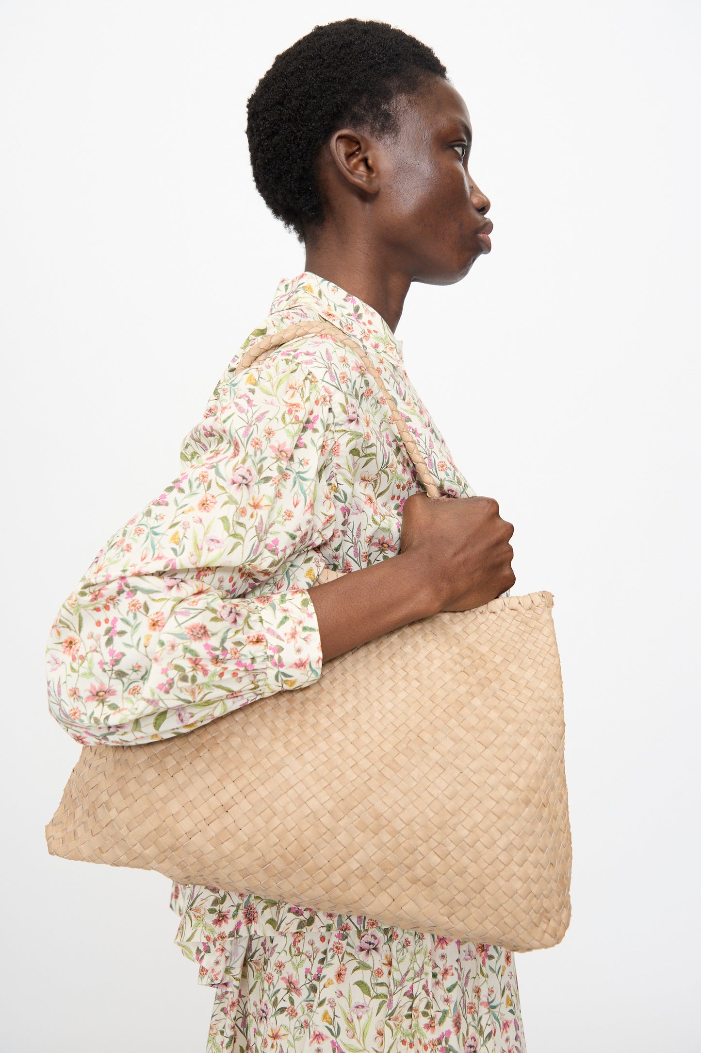 Person in a floral outfit holds the Ichi Antiquités Leather Tote Bag in Greige, featuring braided handles, over their shoulder, shown in profile against a plain background.