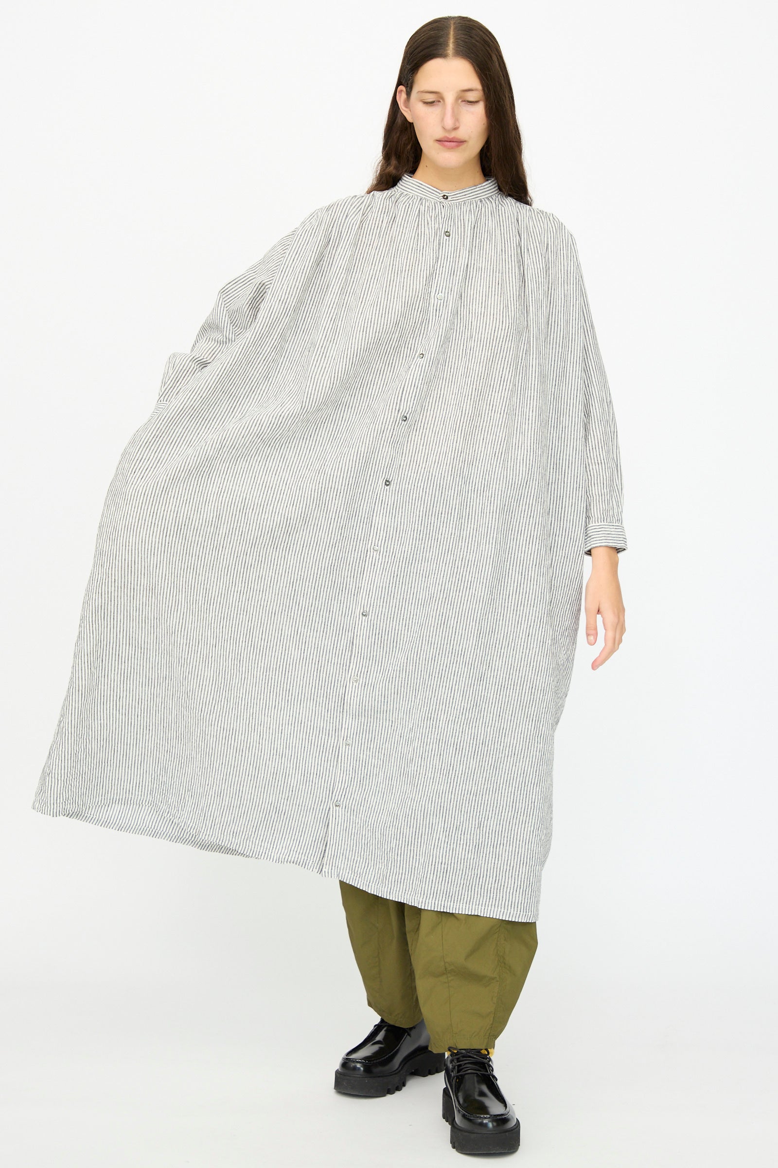 A person models the Ichi Antiquités Linen and Cotton Dress in White Stripe over olive green pants and black shoes, standing against a plain white background.
