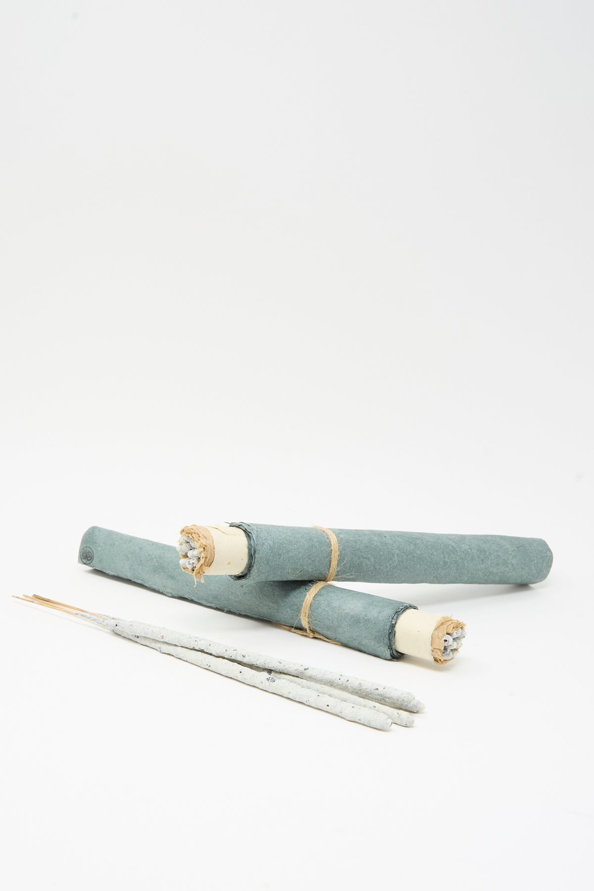 Two grey paper-wrapped Incausa Copal Incense Scrolls tied with string and three thin sticks lying on a white surface.