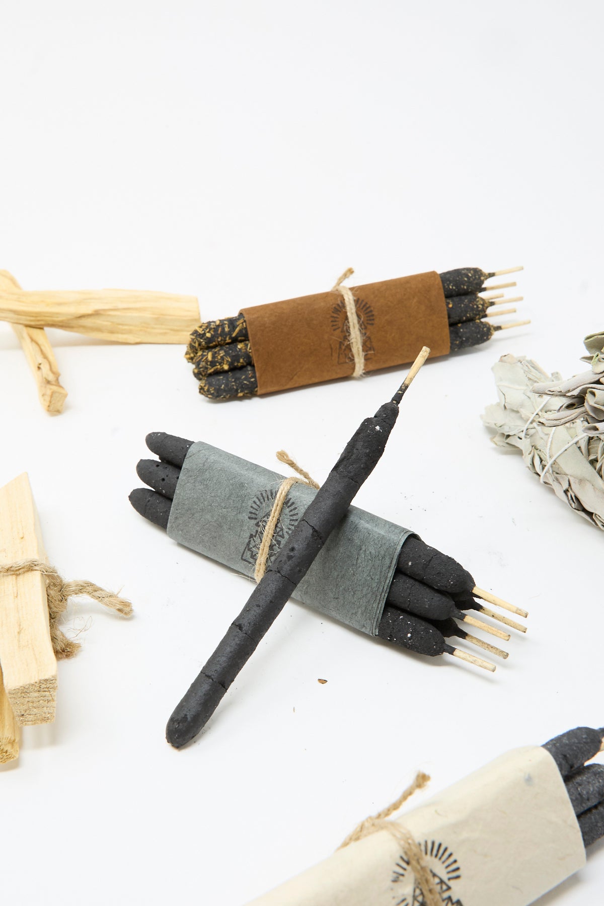 Half Dozen Incense Bundles in White Sage by Incausa, tied with paper bands and arranged with pieces of wood and dried sage on a white surface.