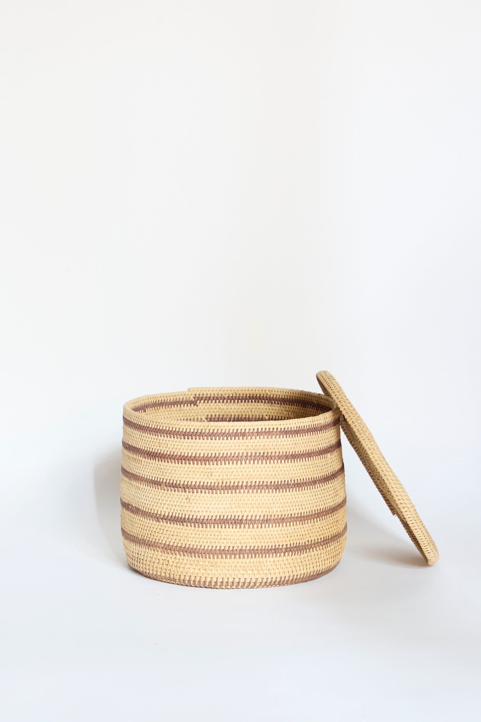 The Motorohina Basket with Yanomami Stripe Painting by Incausa is pictured with its matching lid resting at an angle beside it against a plain white background. This handwoven basket showcases traditional artistry and natural materials, expertly crafted from durable Titica vine in a round shape with distinct brown and beige stripes.