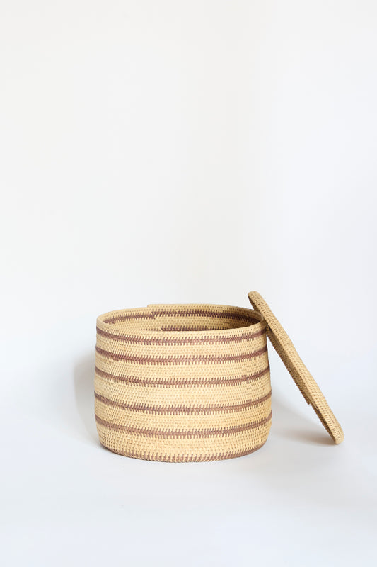 The Motorohina Basket with Yanomami Stripe Painting by Incausa is pictured with its matching lid resting at an angle beside it against a plain white background. This handwoven basket showcases traditional artistry and natural materials, expertly crafted from durable Titica vine in a round shape with distinct brown and beige stripes.