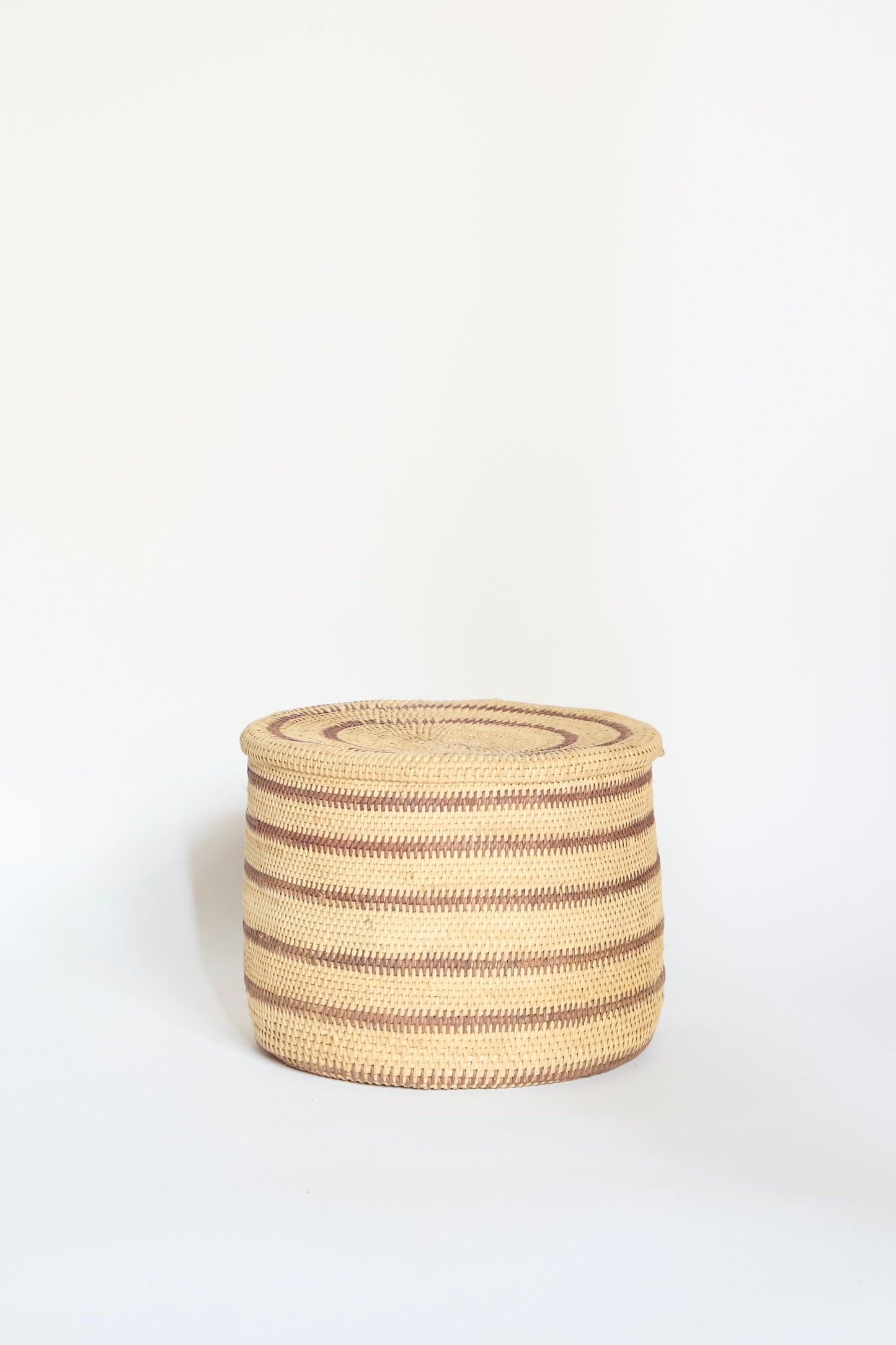 The Motorohina Basket with Yanomami Stripe Painting by Incausa is a round, handwoven basket with a lid, featuring horizontal brown and beige stripes crafted from Titica vine, shown against a plain white background.