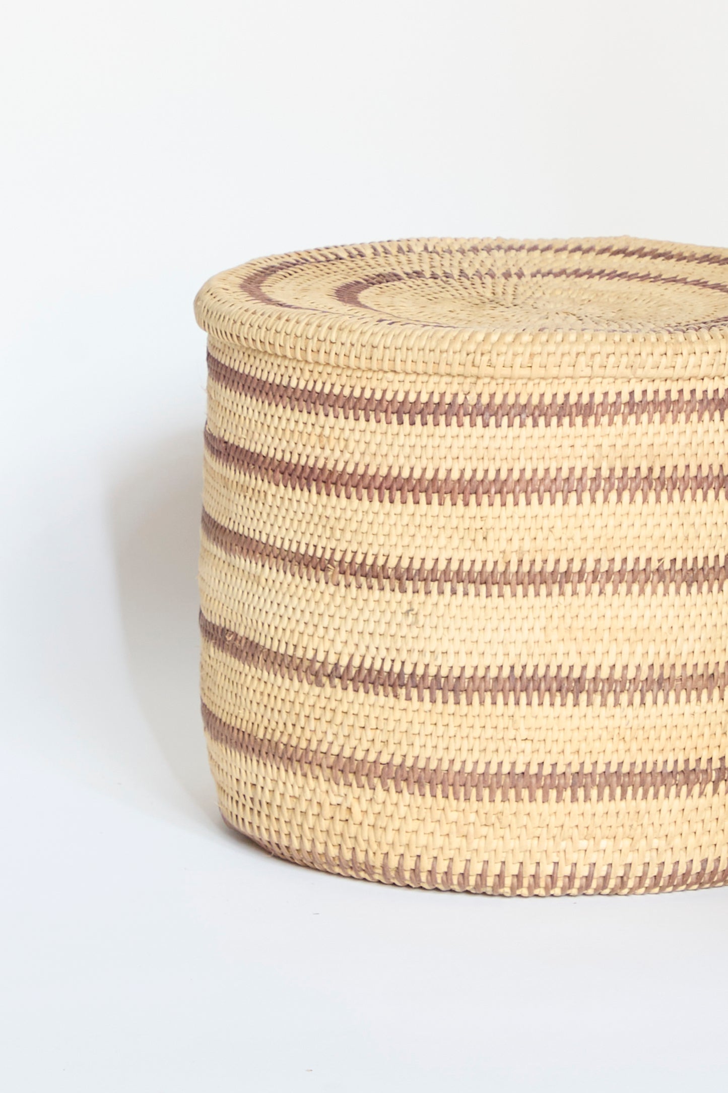 The Motorohina Basket with Yanomami Stripe Painting by Incausa, featuring alternating light and dark horizontal stripes, sits on a plain white background.