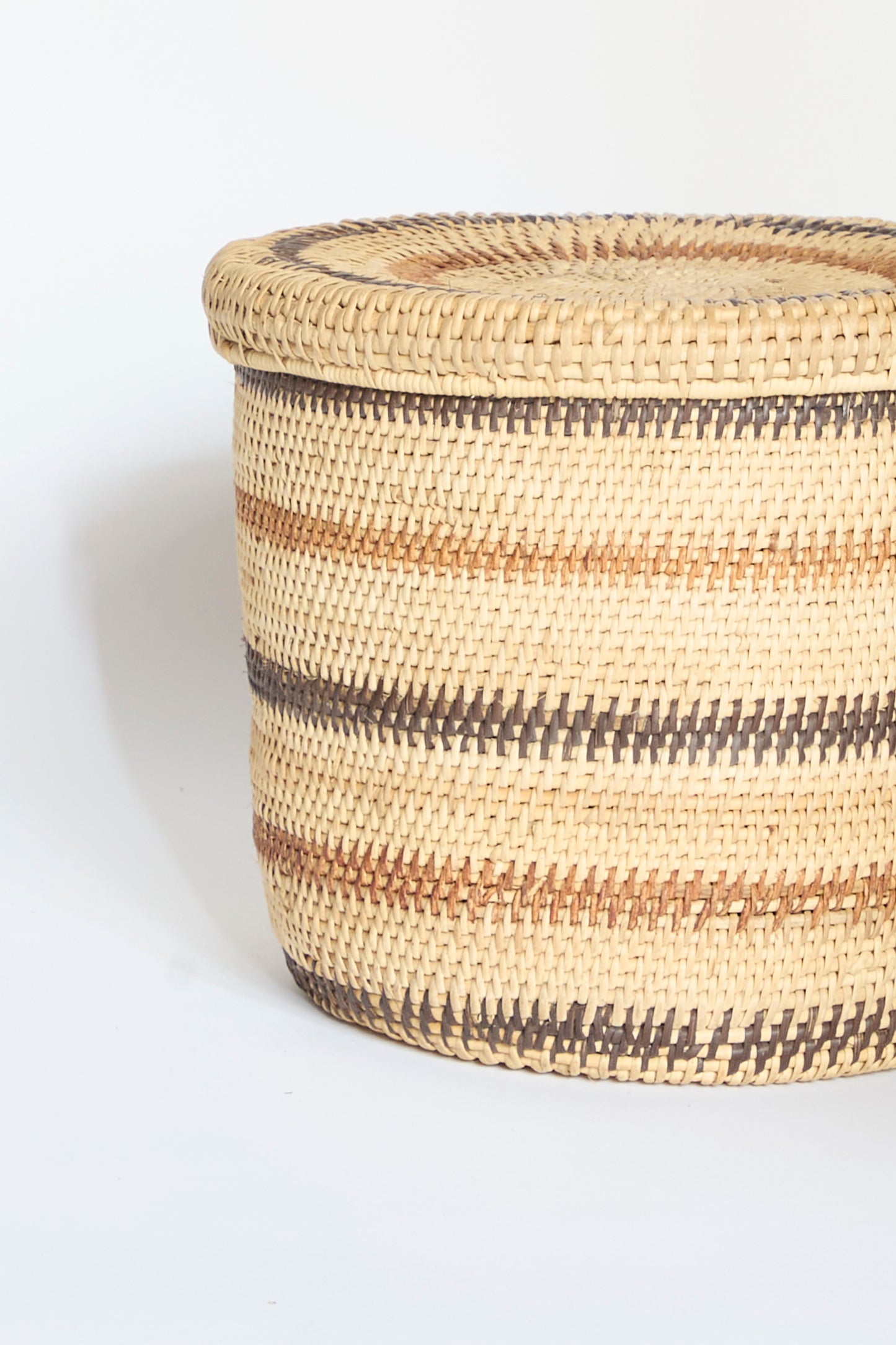A round Motorohina Basket with Yanomami Stripe Painting Mixed by Incausa, handwoven with a lid and featuring alternating natural and dark brown horizontal stripes, set against a plain white background.